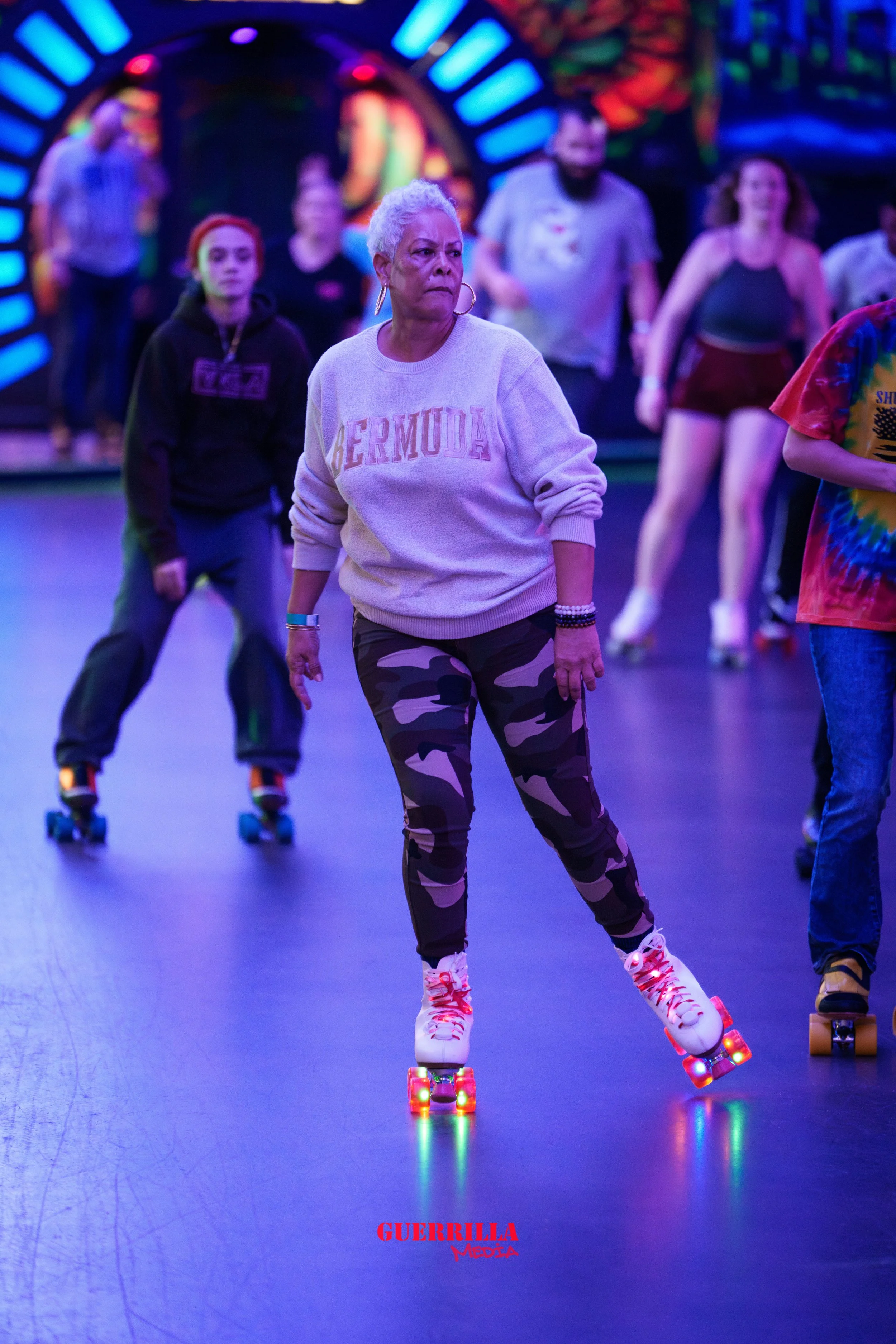 An elderly woman with short white hair roller skating at an indoor skating rink. She is wearing a white sweatshirt with 'BERMUDA' written on it, camouflage pants, and white roller skates with colorful lights. Other people are roller skating in the ba
