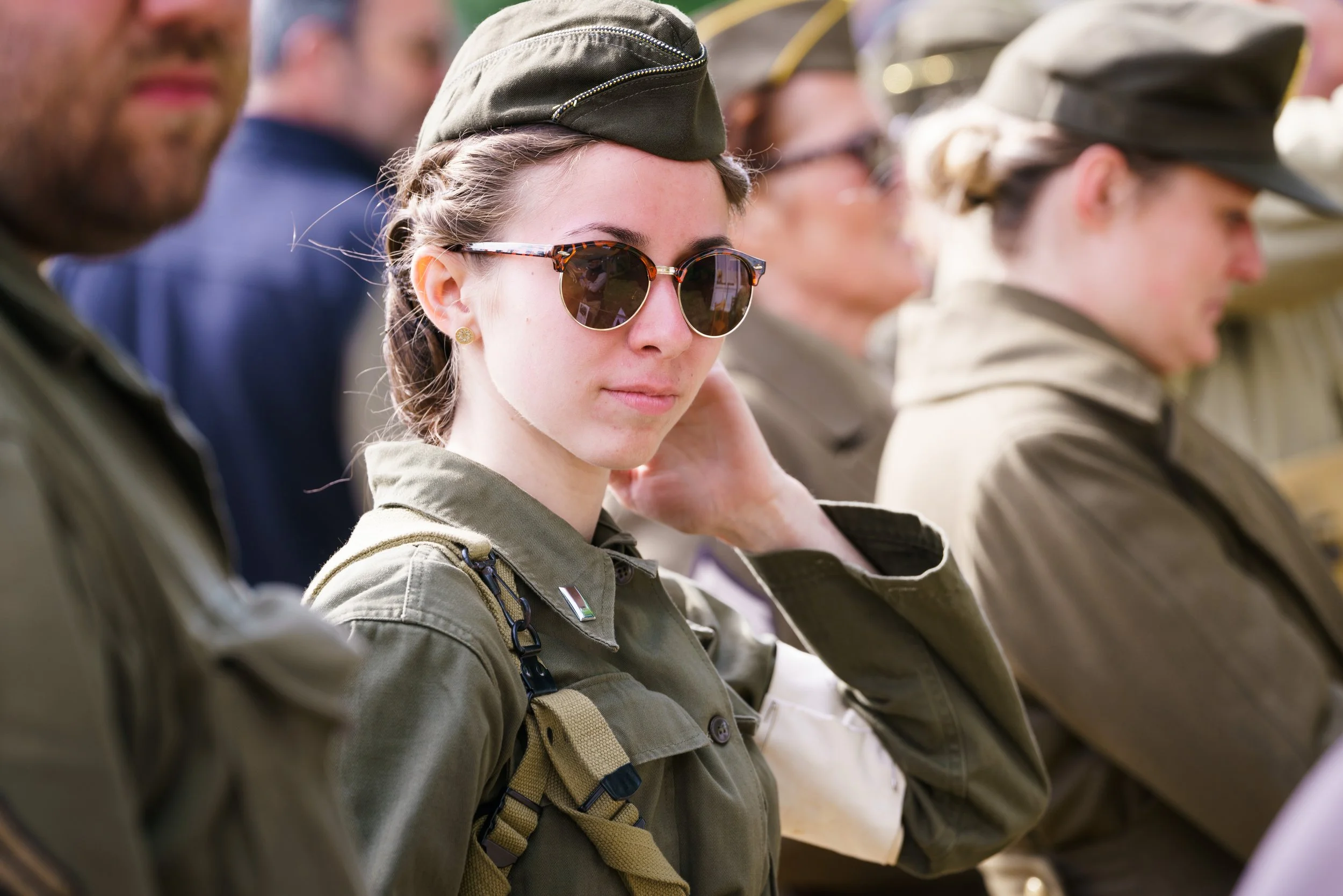 Young woman in military uniform wearing sunglasses, sitting among other soldiers during an outdoor event.