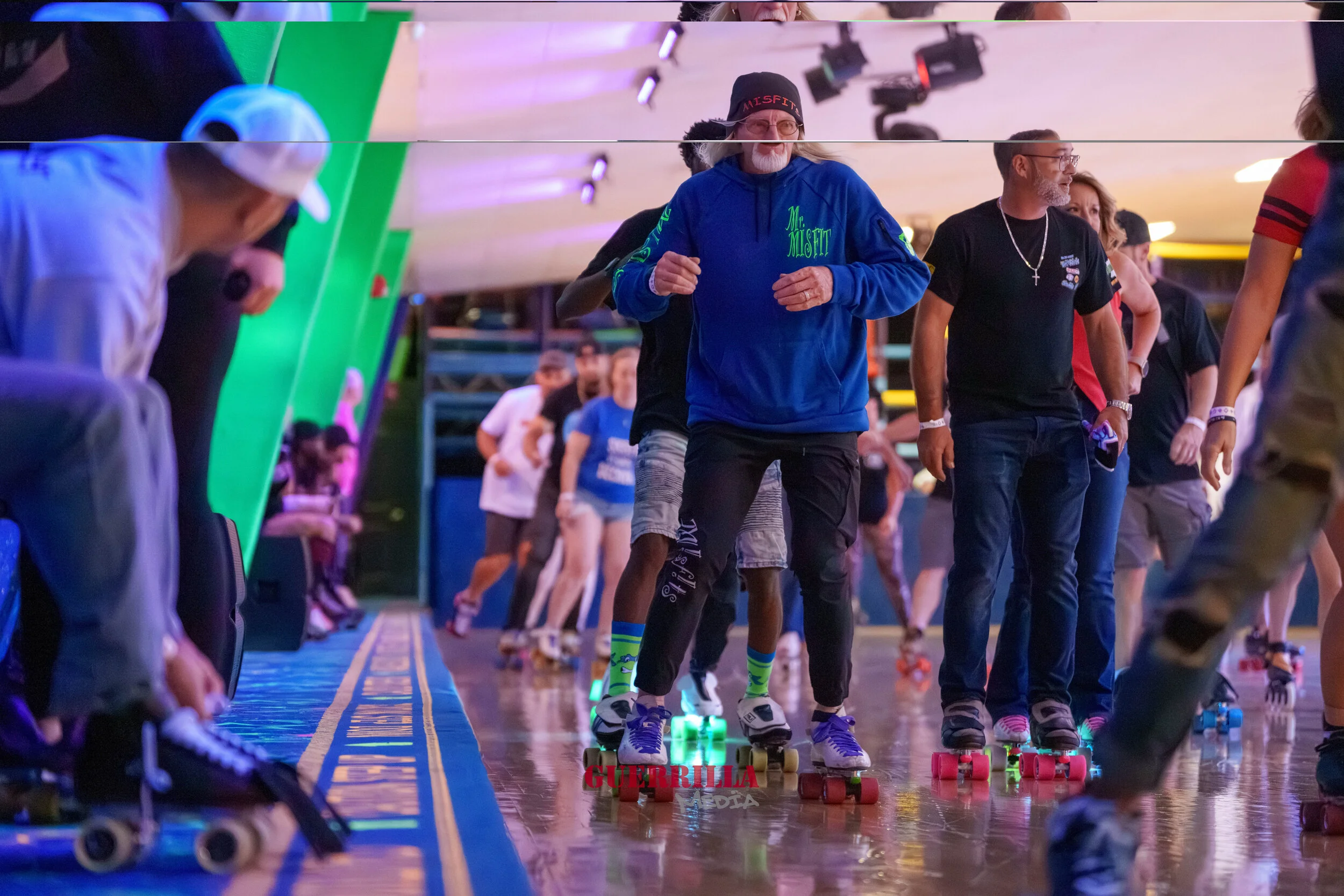 People roller skating indoors, with a man in a blue hoodie in the center and others around him, some wearing casual clothes and some with skate gear.