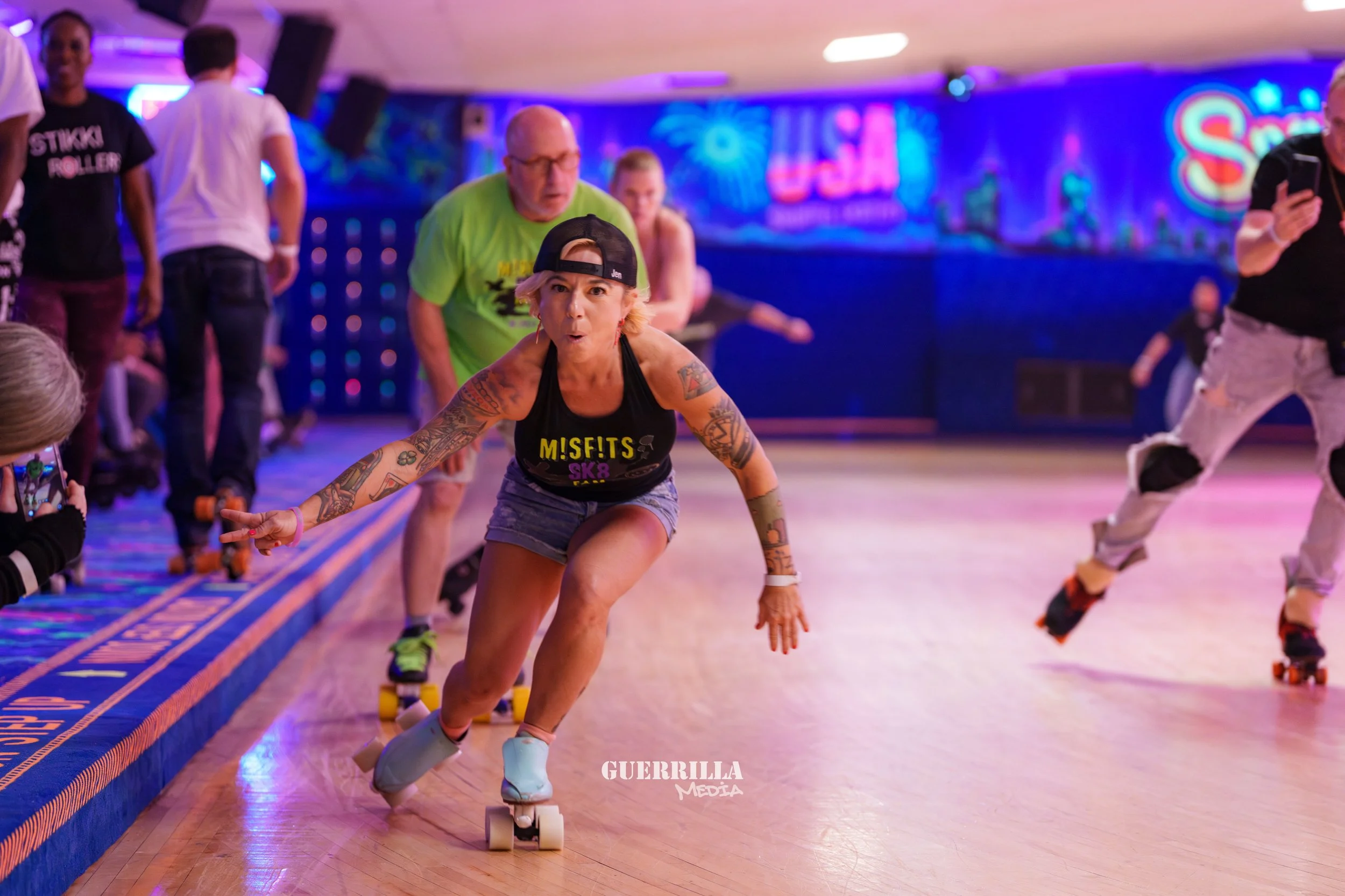 Woman roller skating in an indoor roller rink, wearing a black tank top, denim shorts, and a backward black cap, with tattoos on her arms, making a peace sign and skating forward, with others skating and spectating in the background.