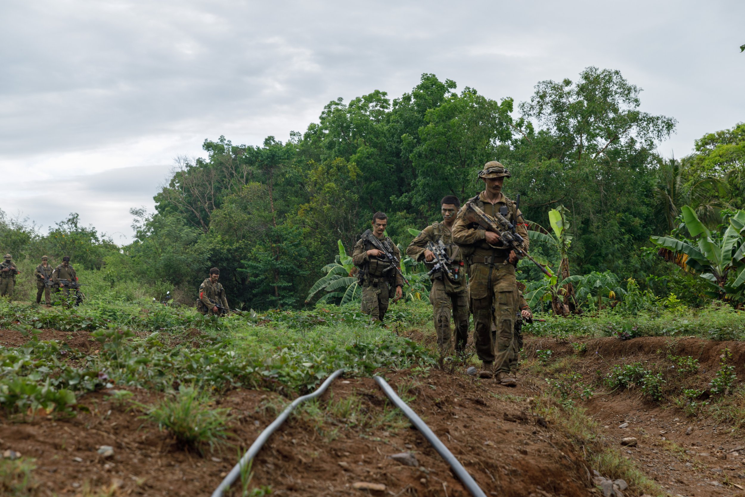 A group of soldiers in camouflage uniforms walking in a line through a green, lush jungle area with trees and banana plants, under cloudy skies.