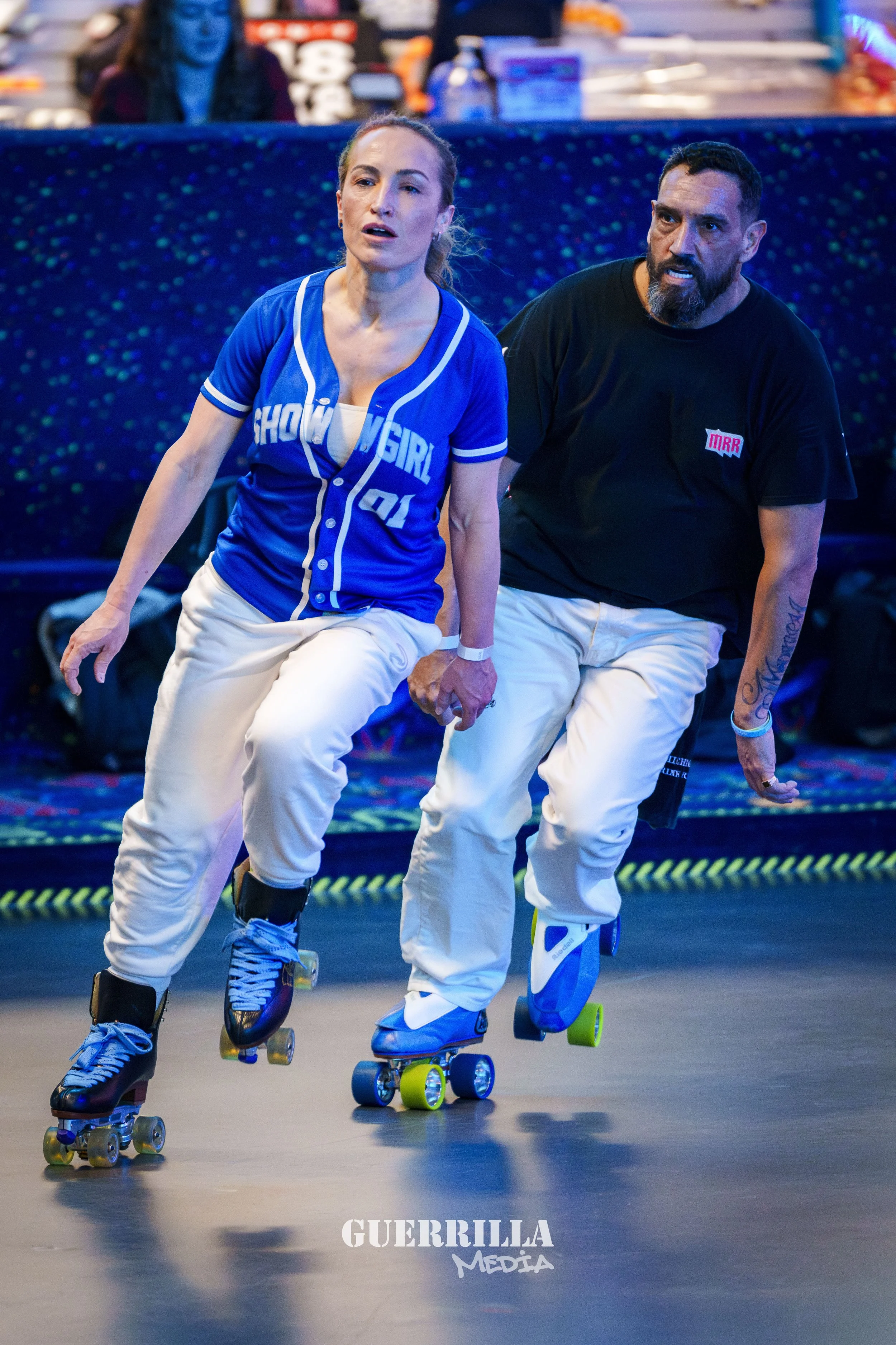 Two people roller skating indoors, one wearing a blue jersey with 'SHOWGIRL' written on it, and the other in a black T-shirt. The woman has her hair tied back, and both are focused on skating.