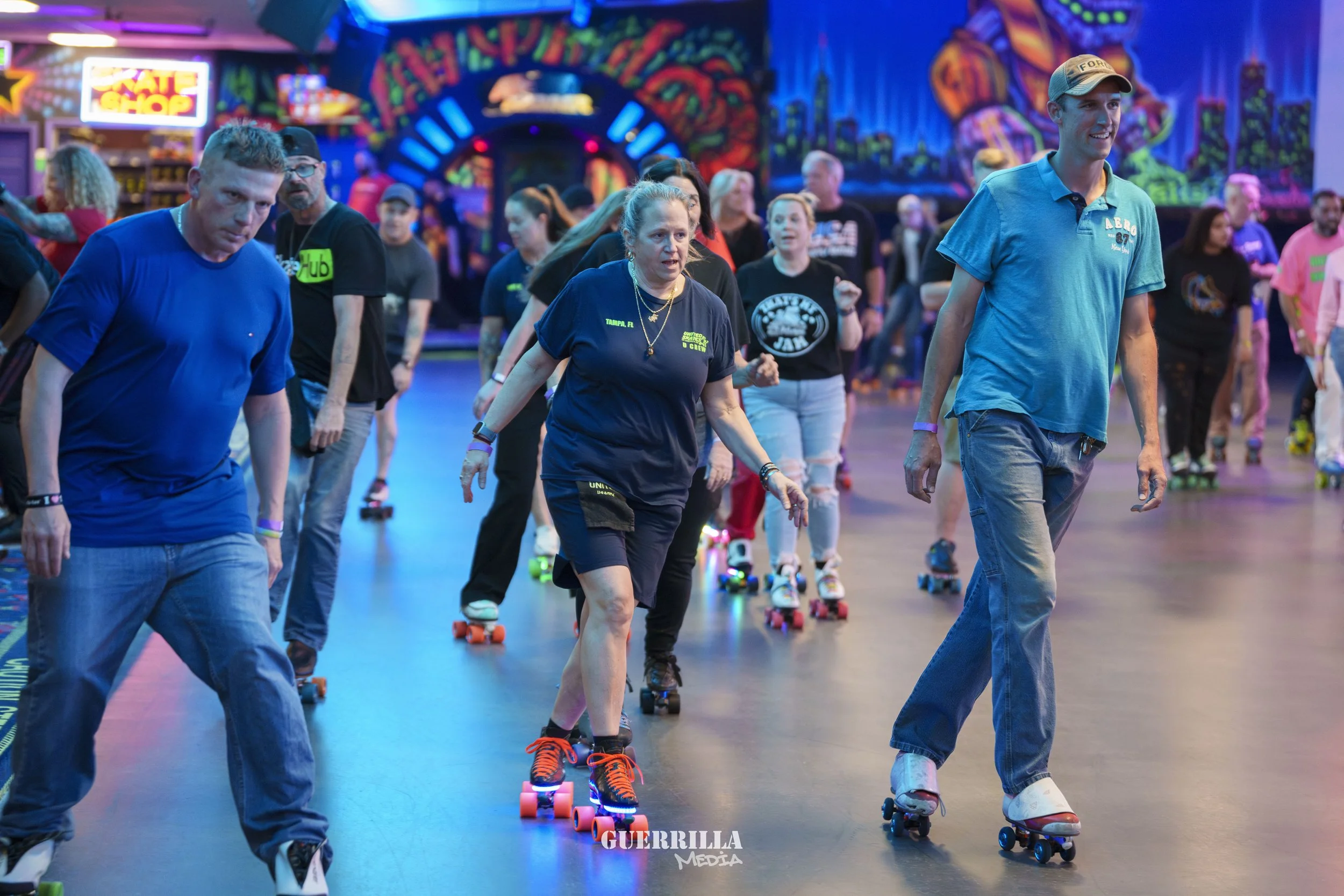 People roller skating inside an amusement park or roller rink with bright, colorful neon lights and painted murals on the walls, creating a lively atmosphere.