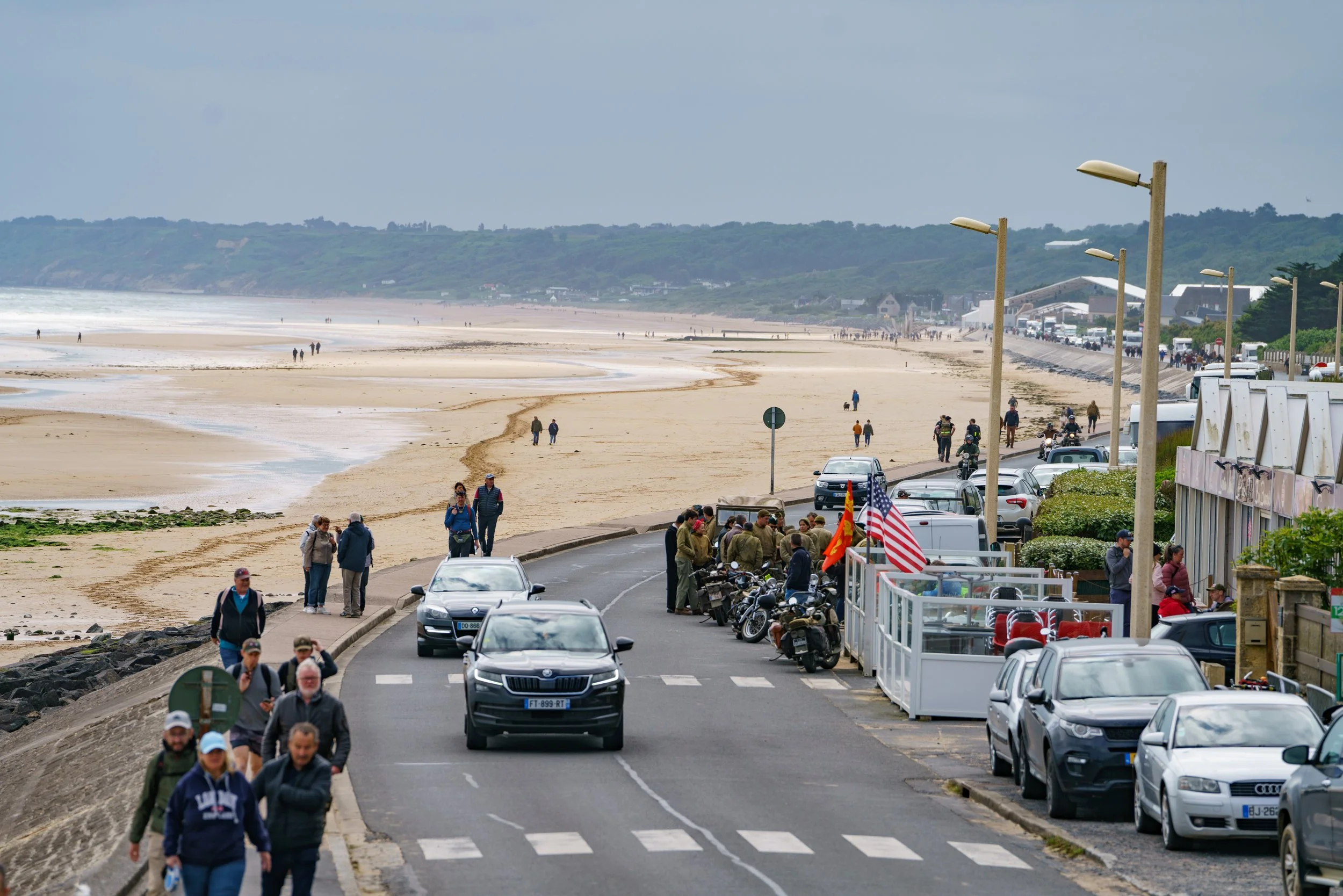 A coastal scene showing a busy street with parked cars and people walking along the sidewalk, leading to a sandy beach and ocean in the background.