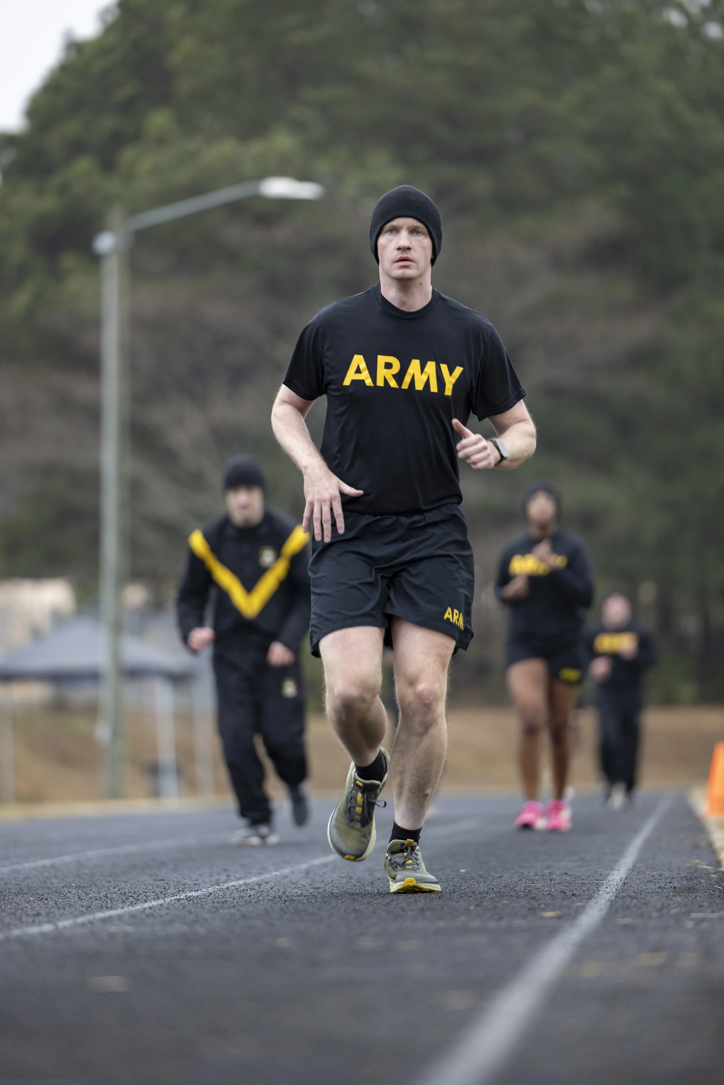 A man wearing a black Army t-shirt and shorts runs on a track with others in running gear behind him, set outdoors with trees and a streetlamp in the background.