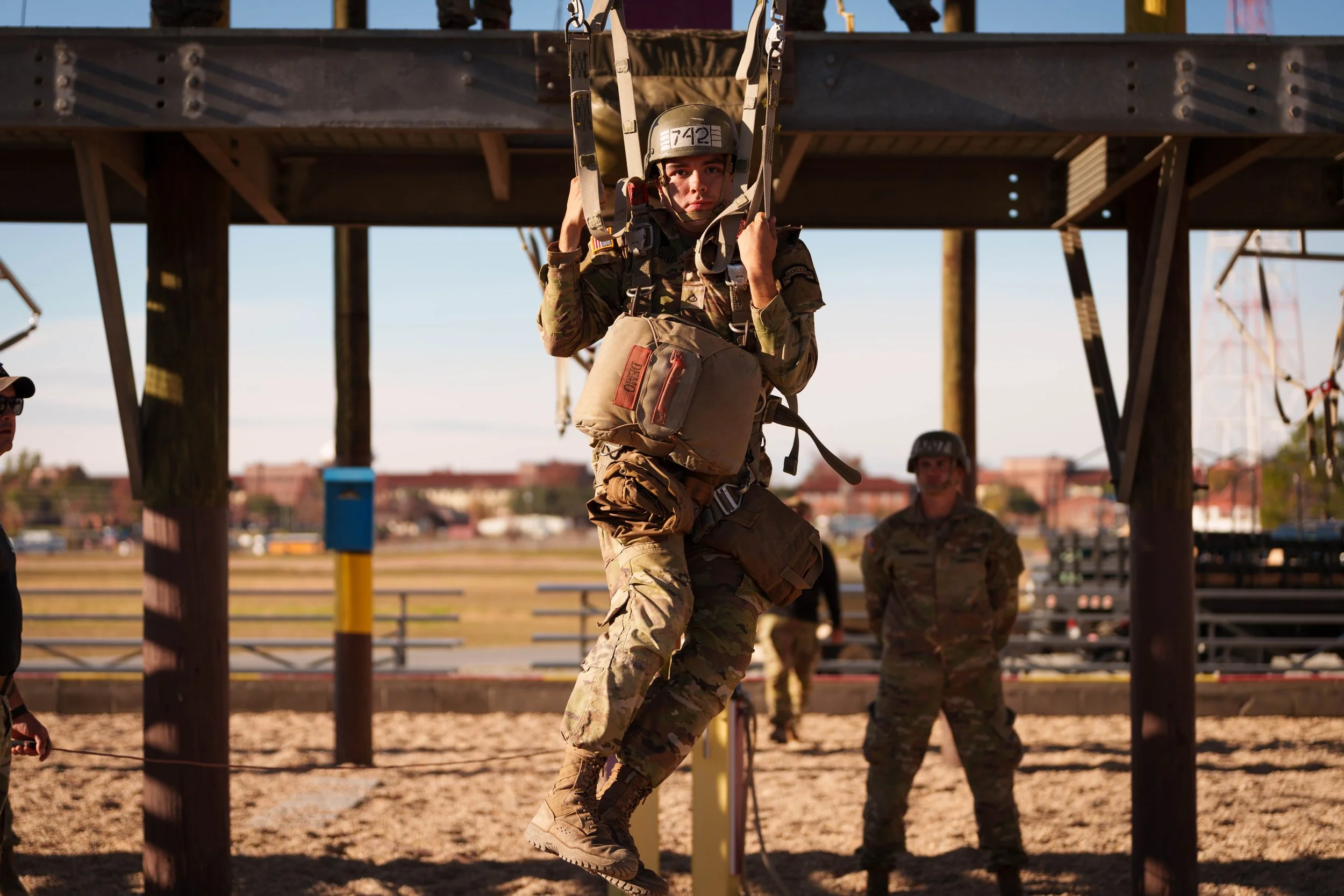 A soldier in camouflage uniform and helmet is jumping from a helicopter harness during a military training exercise, with two other soldiers observing in the background under a structure.