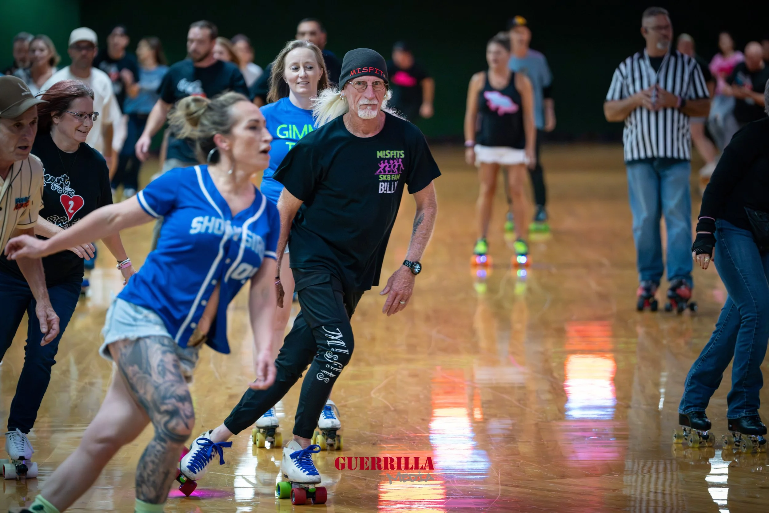 People roller skating indoors, some with vintage or themed clothing, participating in a group skating event or dance, with a wooden floor and a dark background.