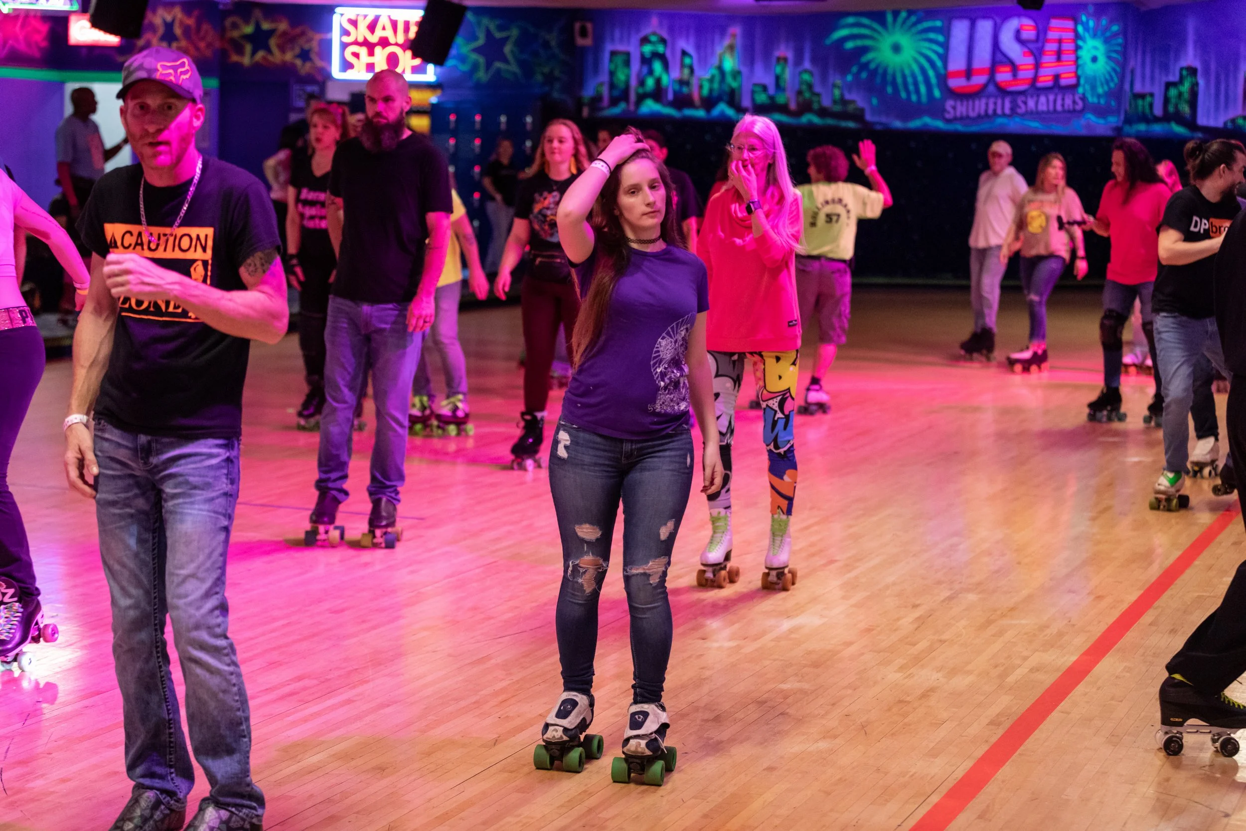People roller skating inside a roller rink with neon lights, colorful mural of a city skyline, and signs that say 'Skate Shop' and 'USA Shuffle Skaters'.
