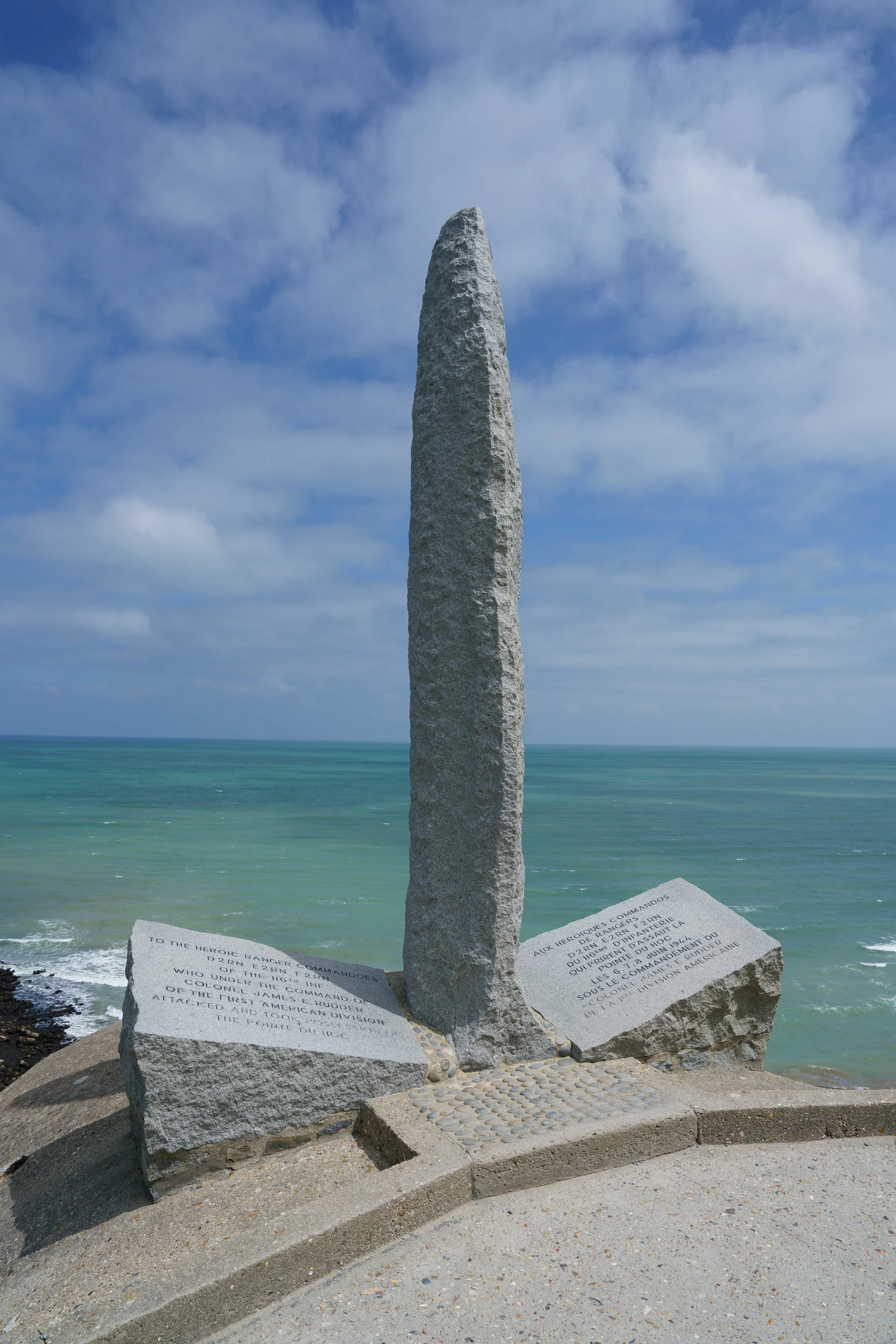 A stone monument resembling a sword, with two inscribed stone plaques at its base, located by the ocean under a partly cloudy sky.