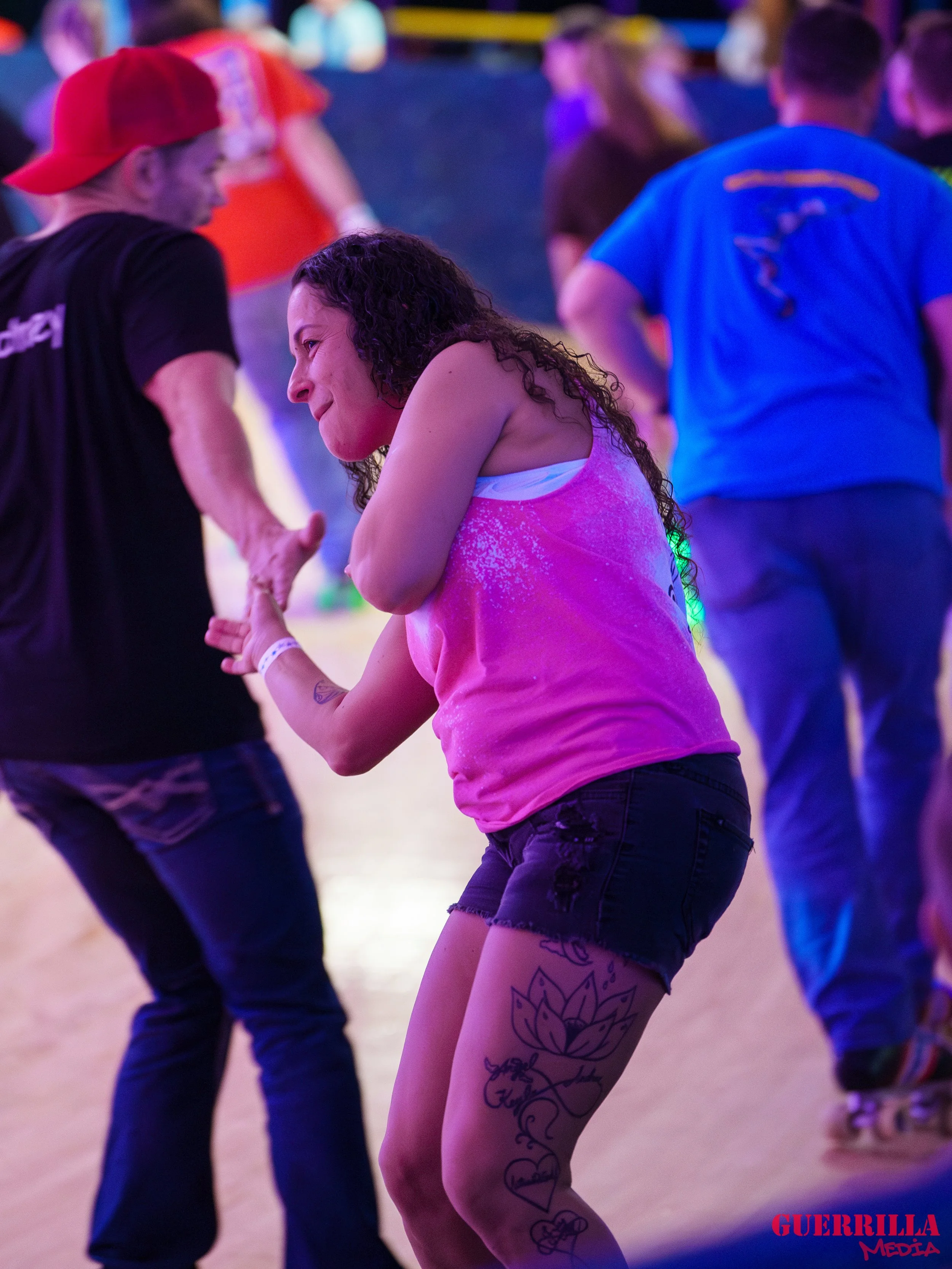 A woman with a tattoo on her thigh, wearing a pink tank top and black shorts, dancing and smiling at a lively indoor event with colorful lighting and other people in the background.