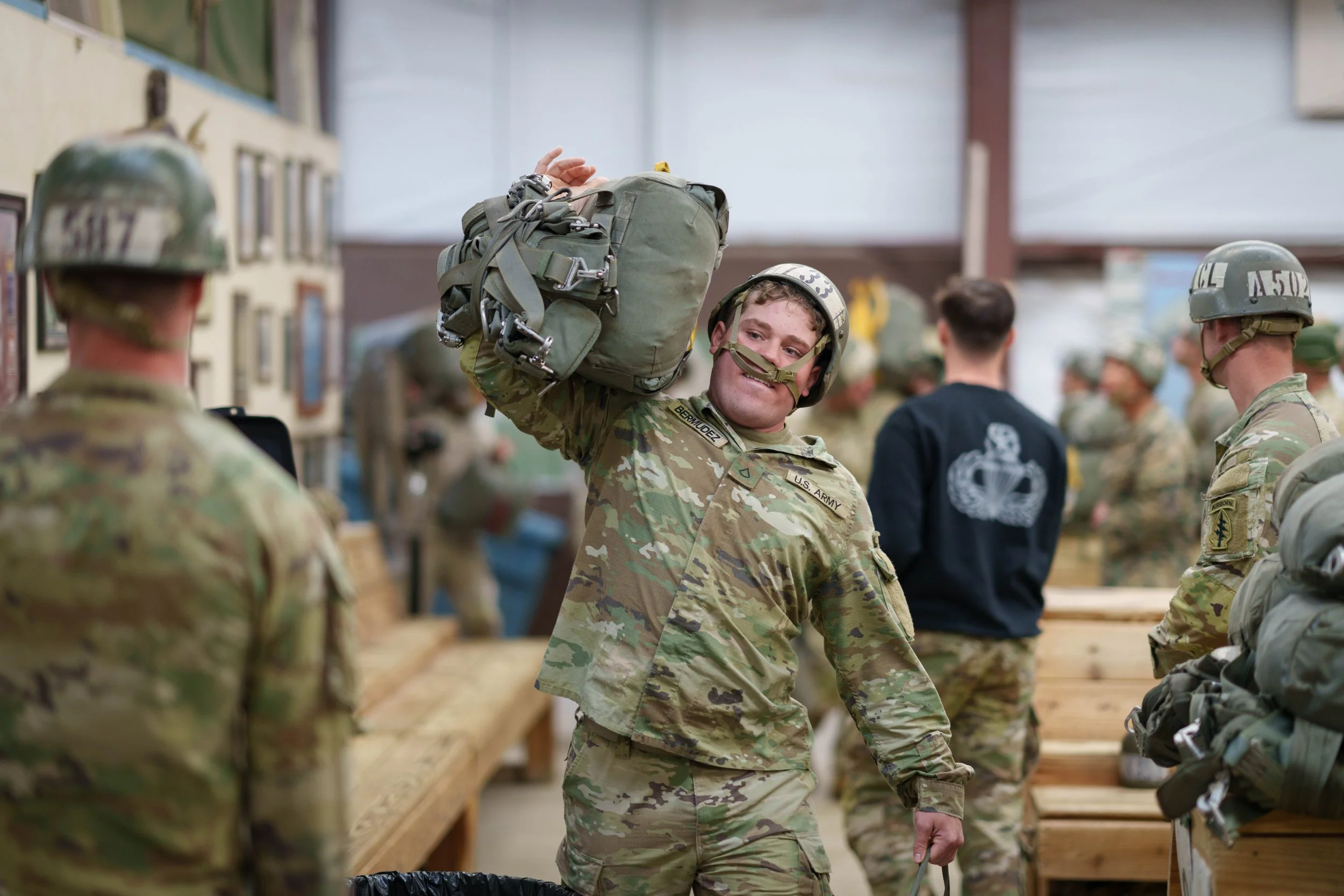 A soldier in camouflage uniform smiling and carrying a backpack, surrounded by other soldiers in a military setting.