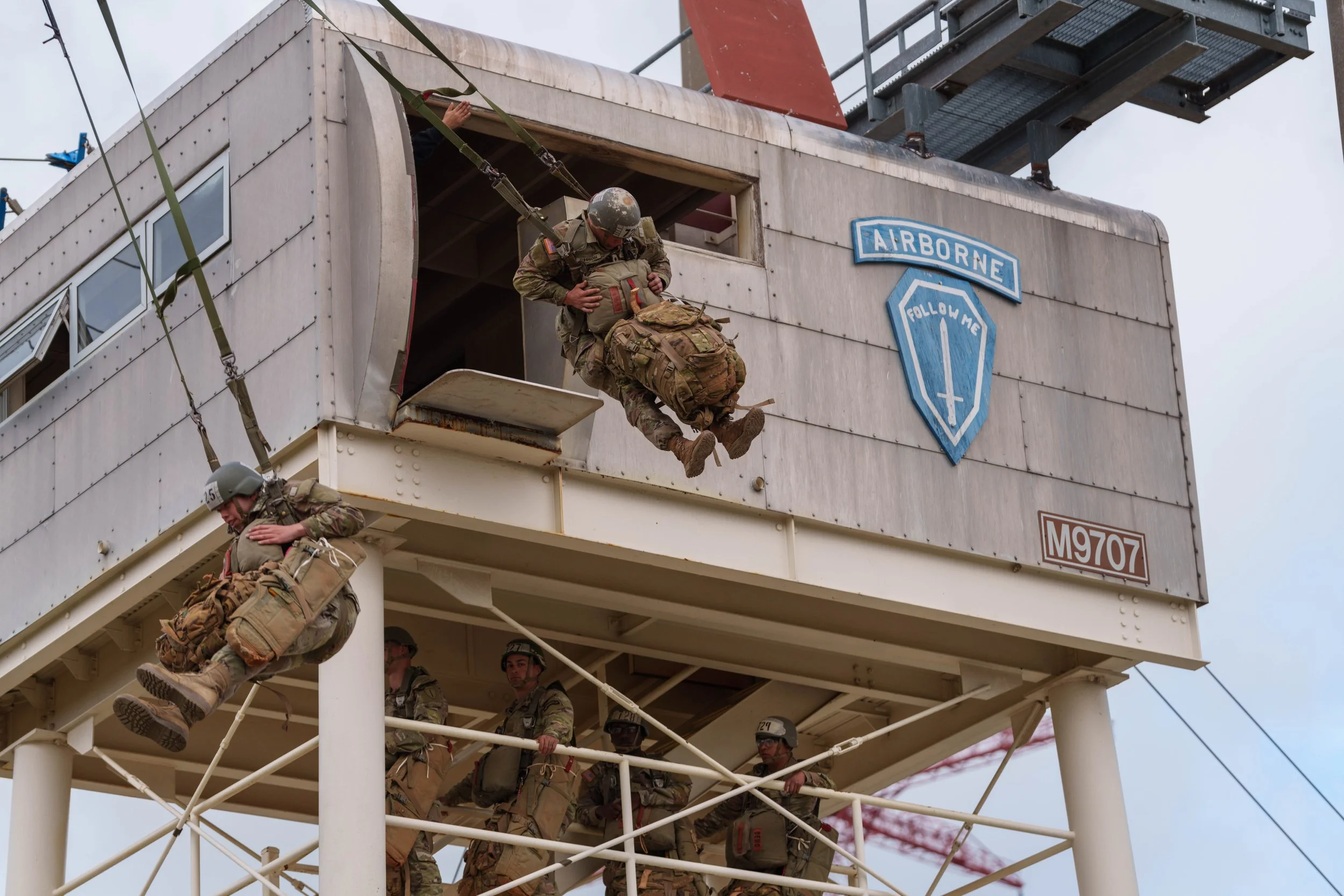 Military personnel involved in a helicopter rescue training exercise, with one soldier being hoisted out of a tower and others on the platform below.