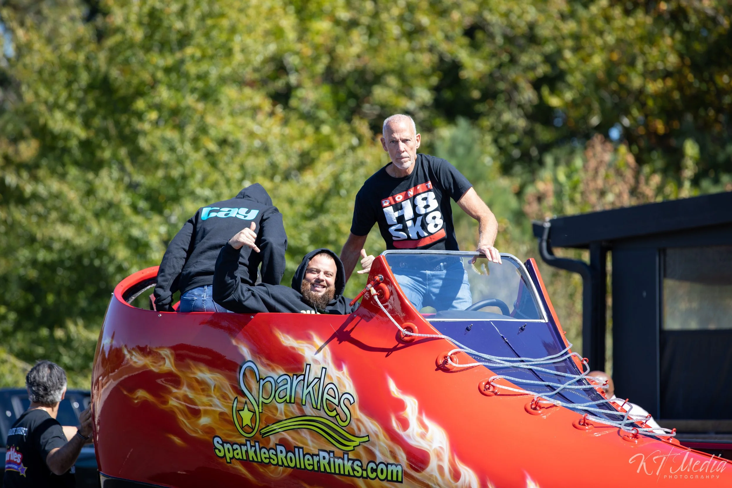 Two men sitting in a red roller coaster car with flame graphics, one giving a thumbs-up and smiling, the other standing behind, on a roller coaster ride at an outdoor amusement park with trees in the background.