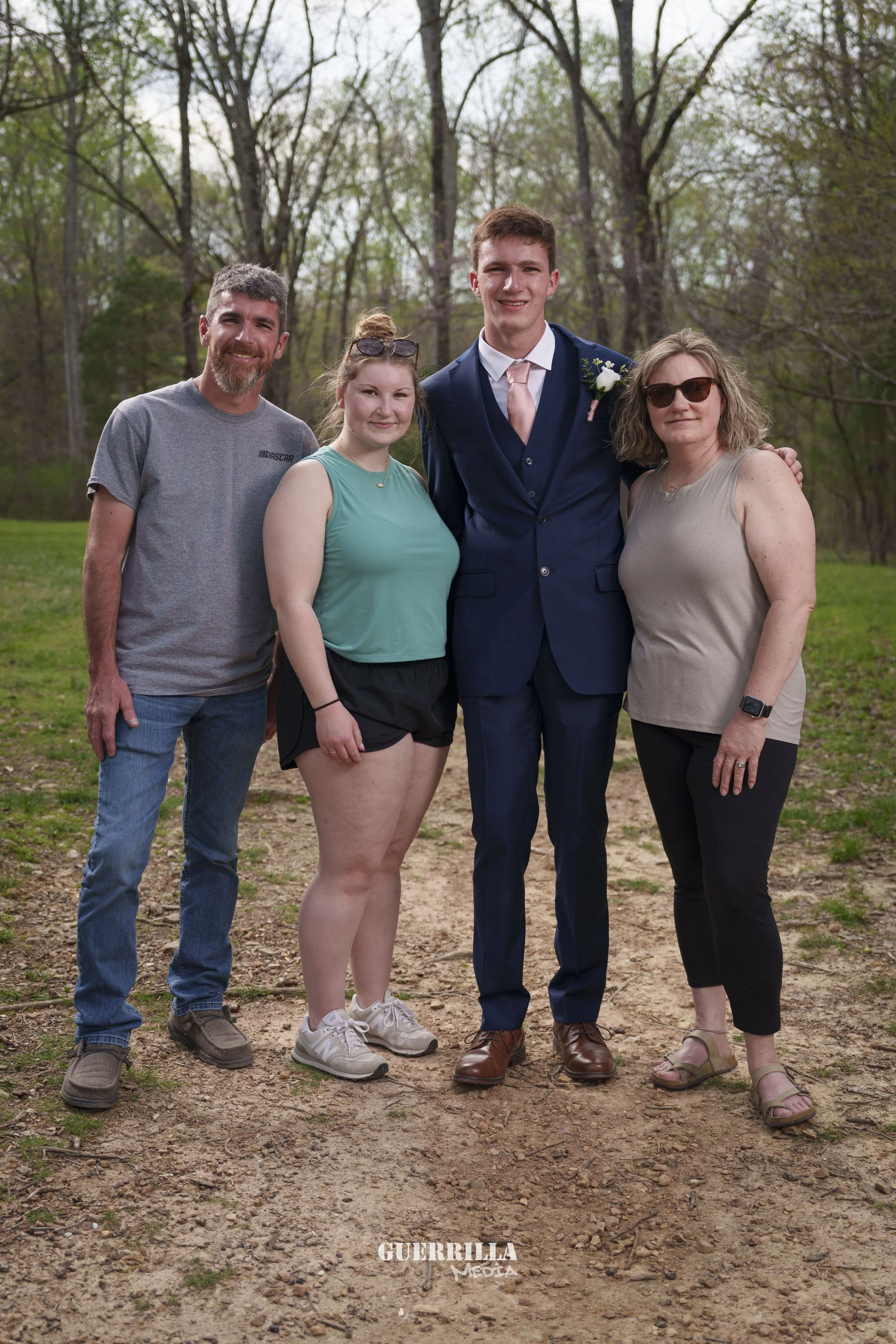A family of four stands outdoors on a dirt path with trees and greenery in the background, posing for a photo. The father and mother stand on either side of a young man dressed in a navy blue suit, pink tie, and white shirt, who appears to be celebra