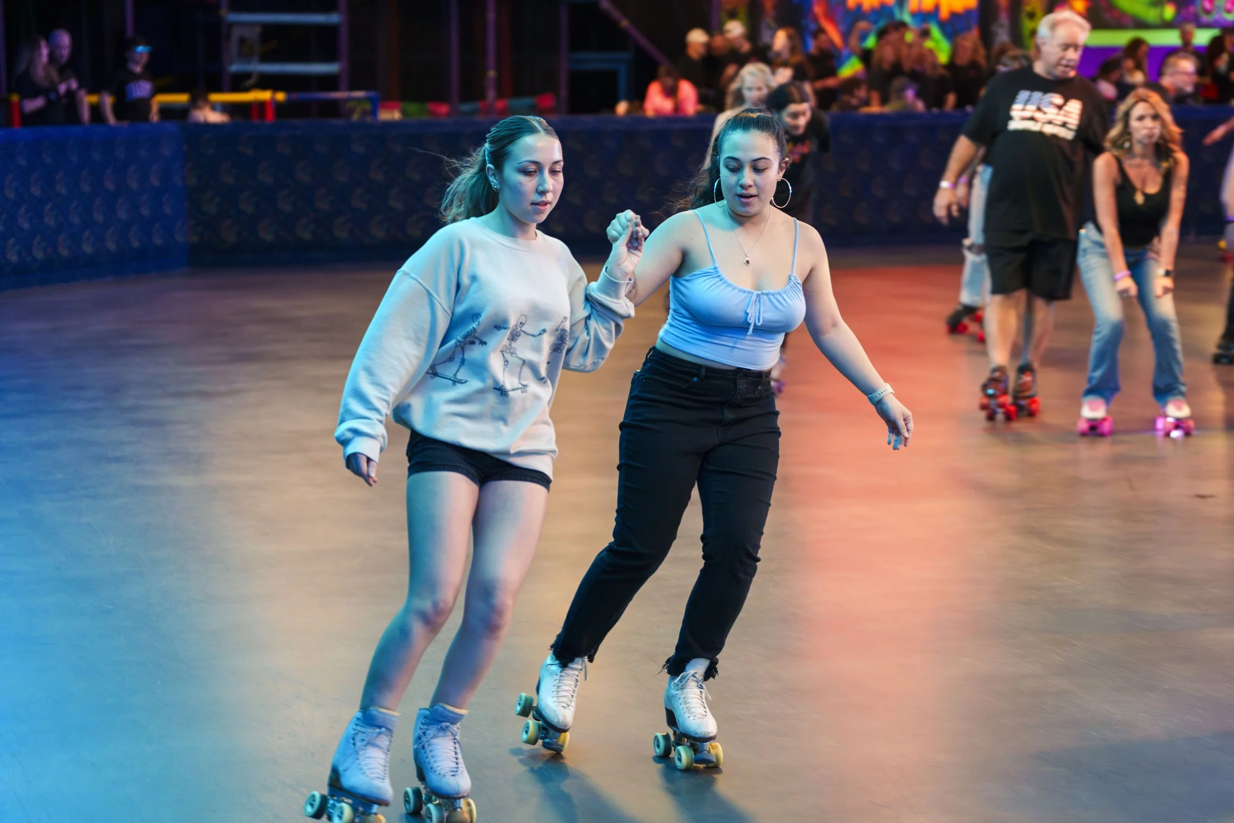 Two young women roller skating together at an indoor roller rink, with other skaters and colorful neon lights in the background.