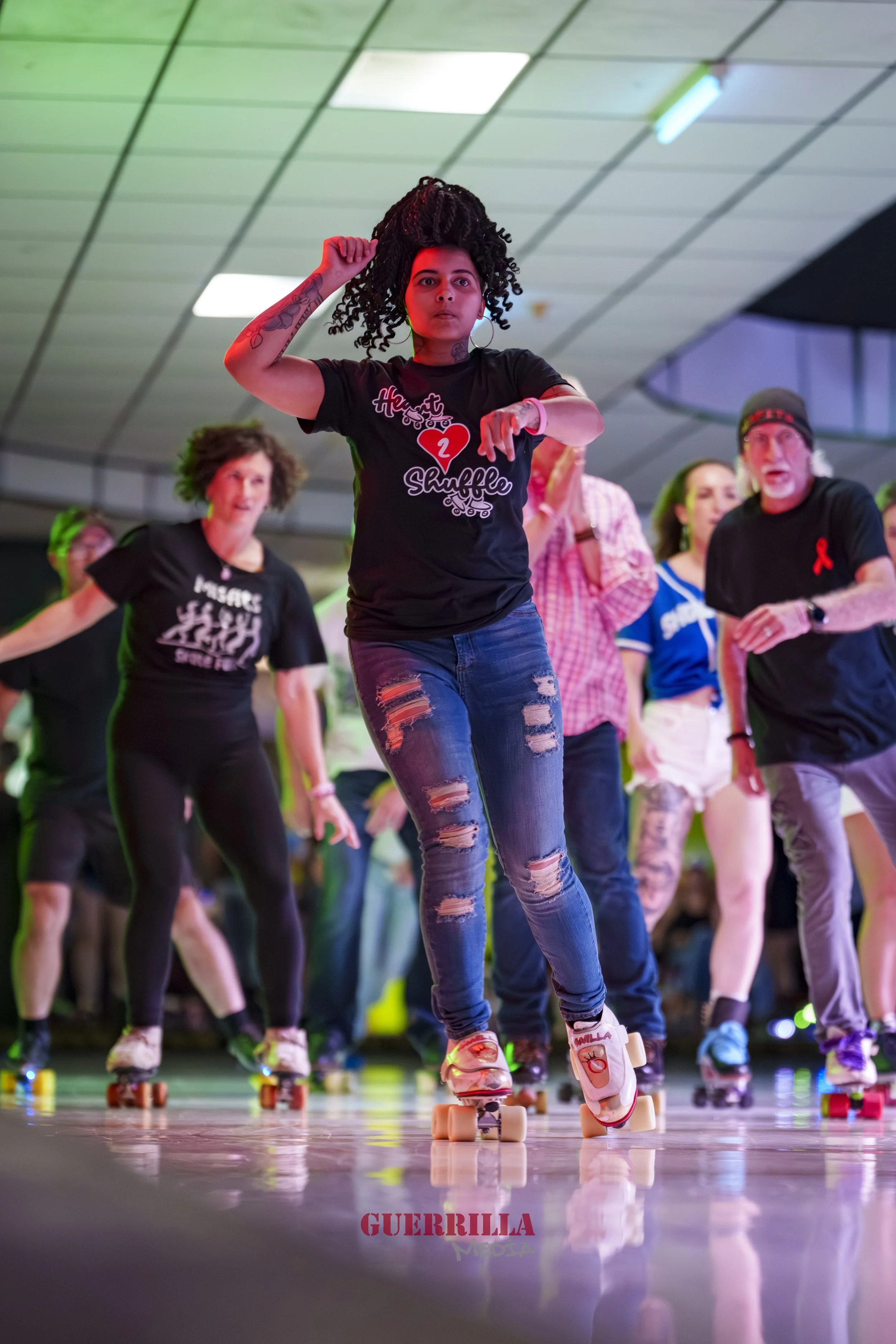 A young woman roller skating indoors with a group of people. She has curly hair, wears a black t-shirt with 'Heart Shuffle,' ripped jeans, and is in a dance or skating pose. Other skaters and spectators are visible in the background.