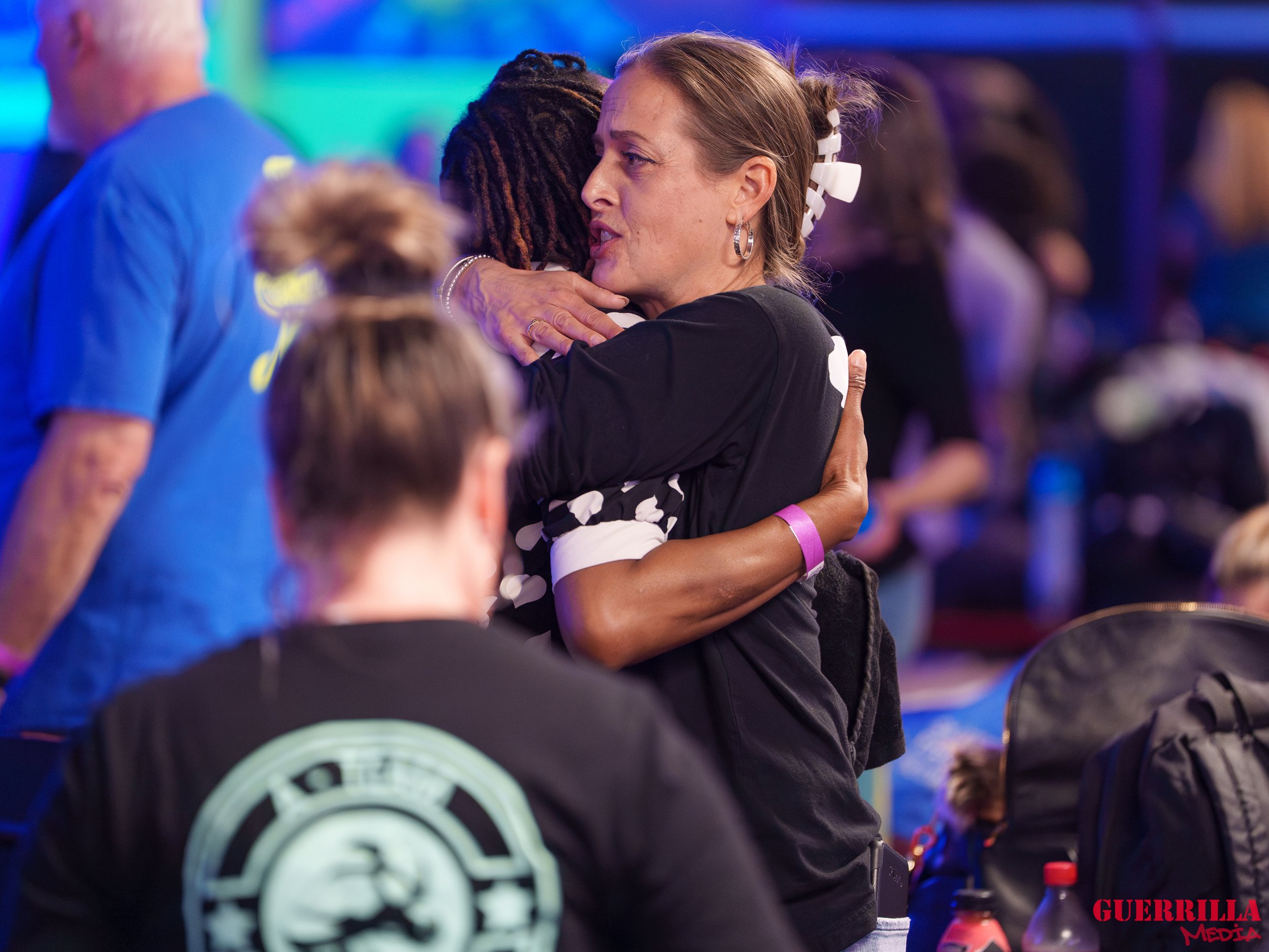 Two women are hugging each other warmly, with one appearing emotional, in a crowded indoor setting with colorful lighting, surrounded by other people and various items like backpacks and bottles.