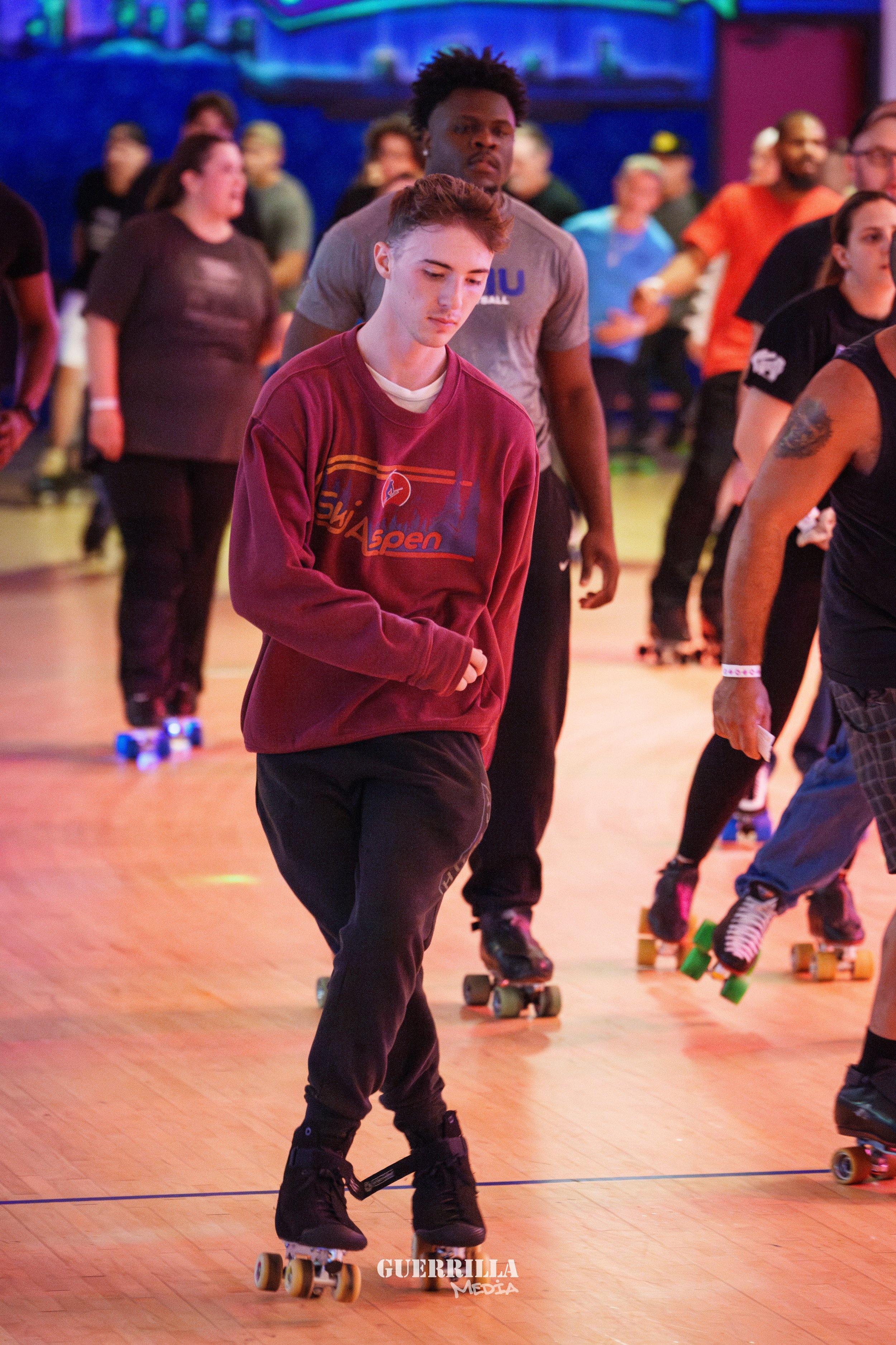 Young man roller skating at an indoor event, surrounded by other skaters and spectators.