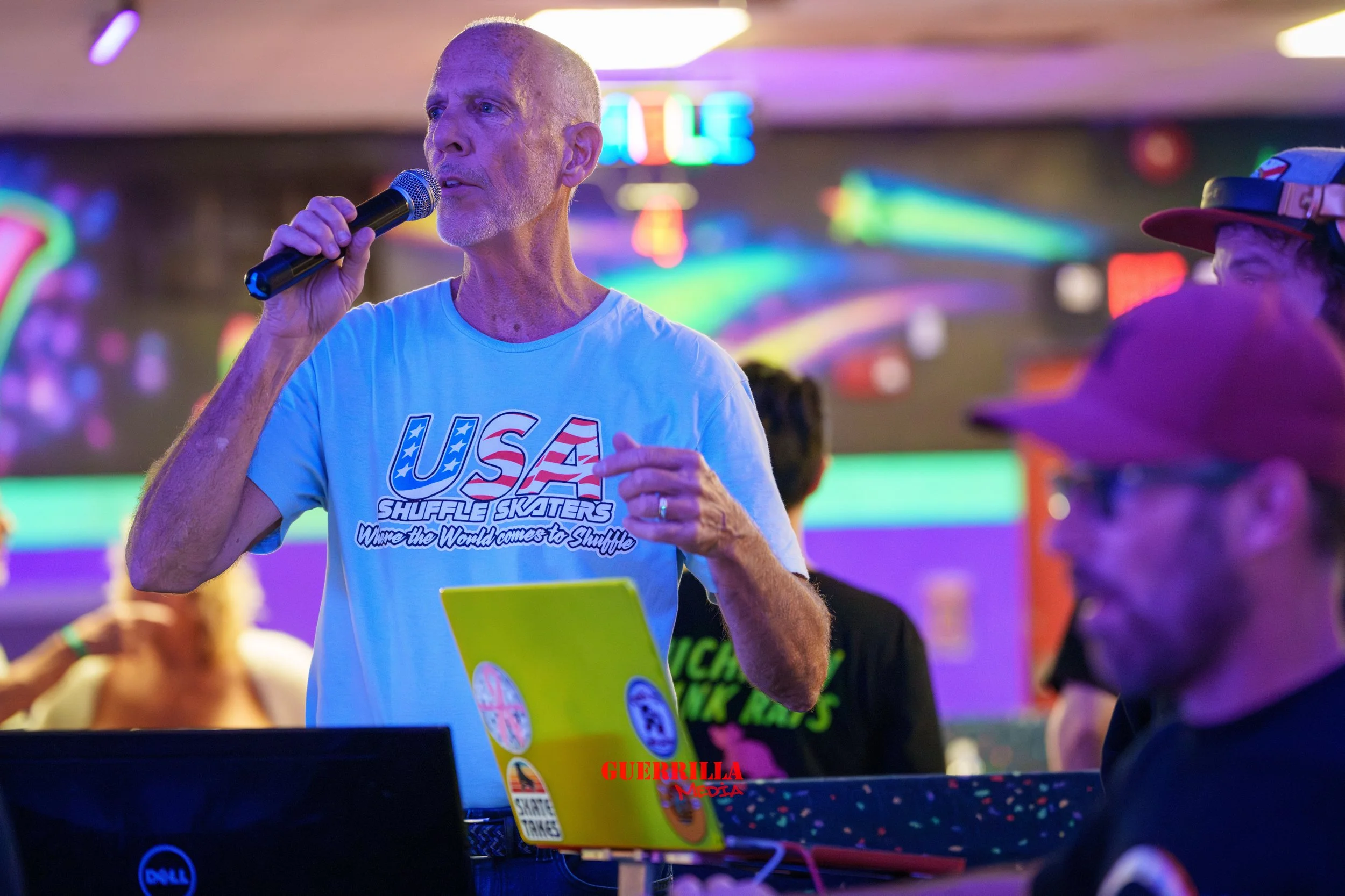 An older man with a microphone in his right hand speaking at an event, wearing a light blue T-shirt with the words "USA Shuffle Skaters" on it, surrounded by people, with neon lights in the background.