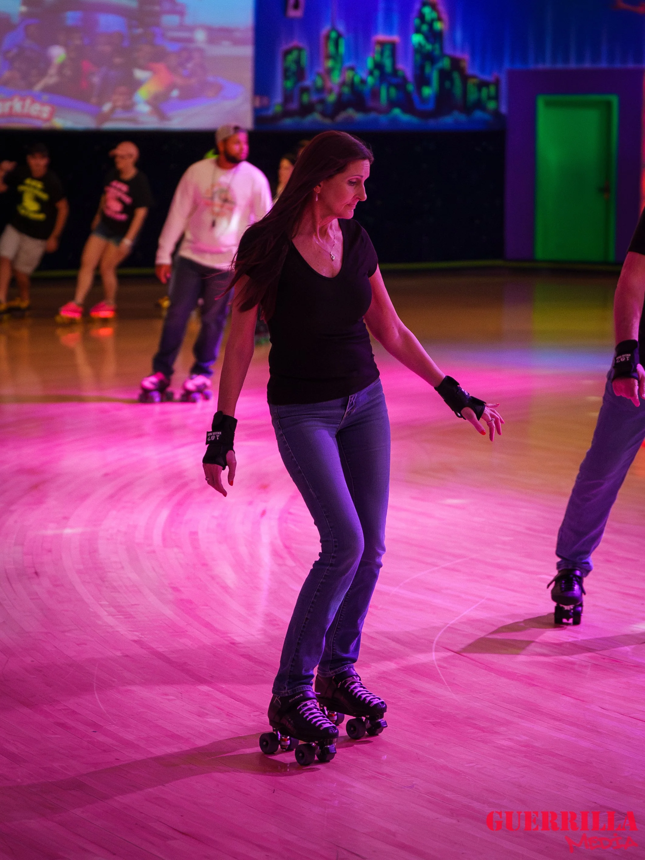 Woman roller skating on a wooden floor under pink and purple lighting at an indoor roller rink, with colorful graffiti-style cityscape on the wall in the background.