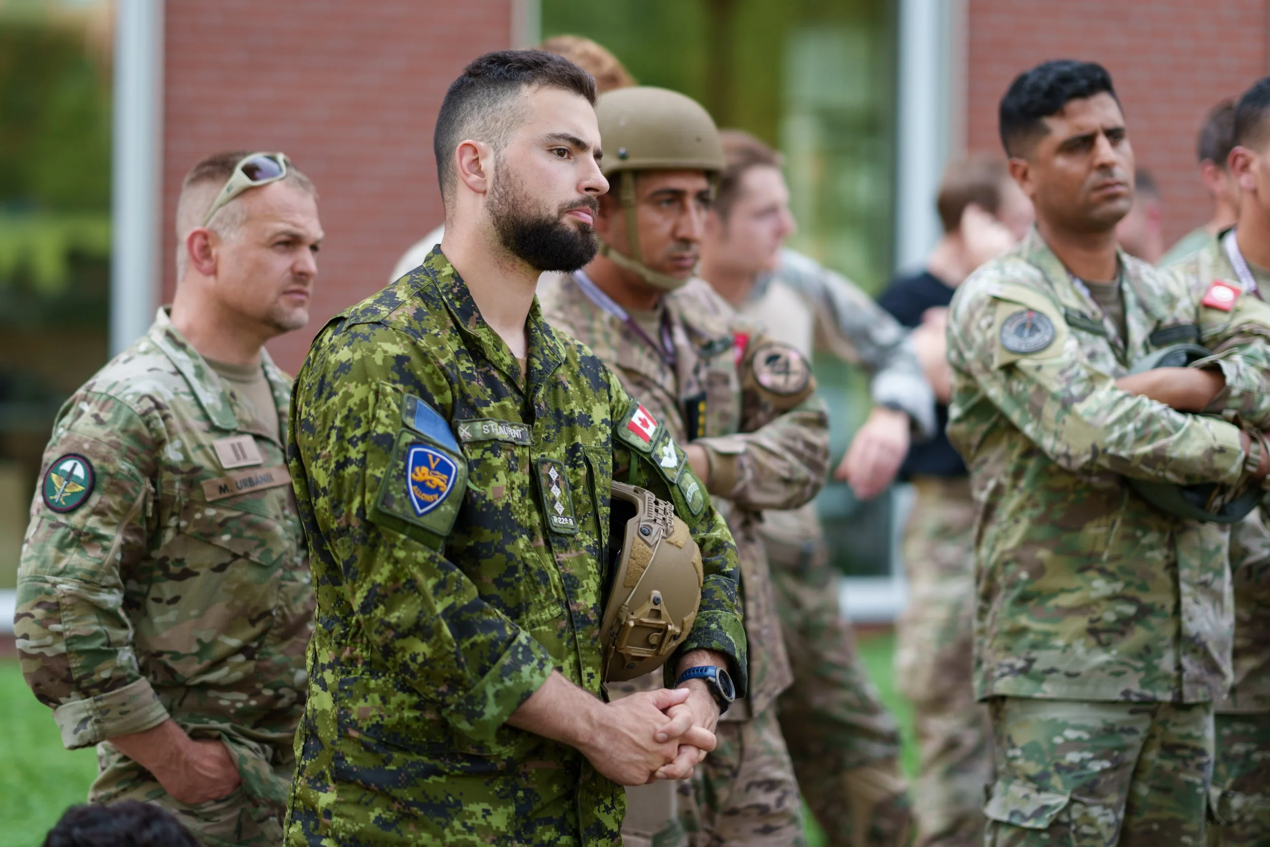 Group of soldiers in uniform, standing outdoors with serious expressions, some with arms crossed or hands clasped.