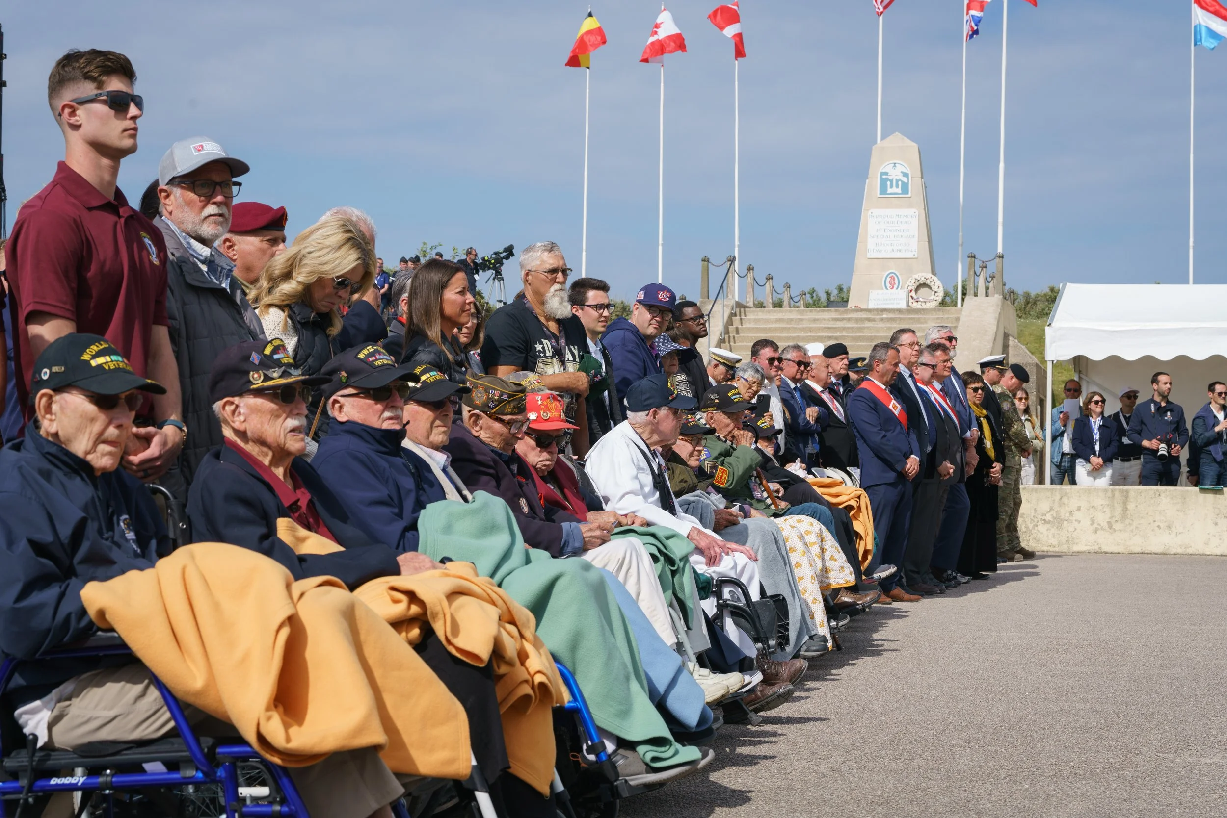 A crowd gathered at a memorial service, including elderly veterans in wheelchairs and younger attendees standing behind them. The event takes place outdoors under a partly cloudy sky, with flags flying on tall poles in the background and a memorial m