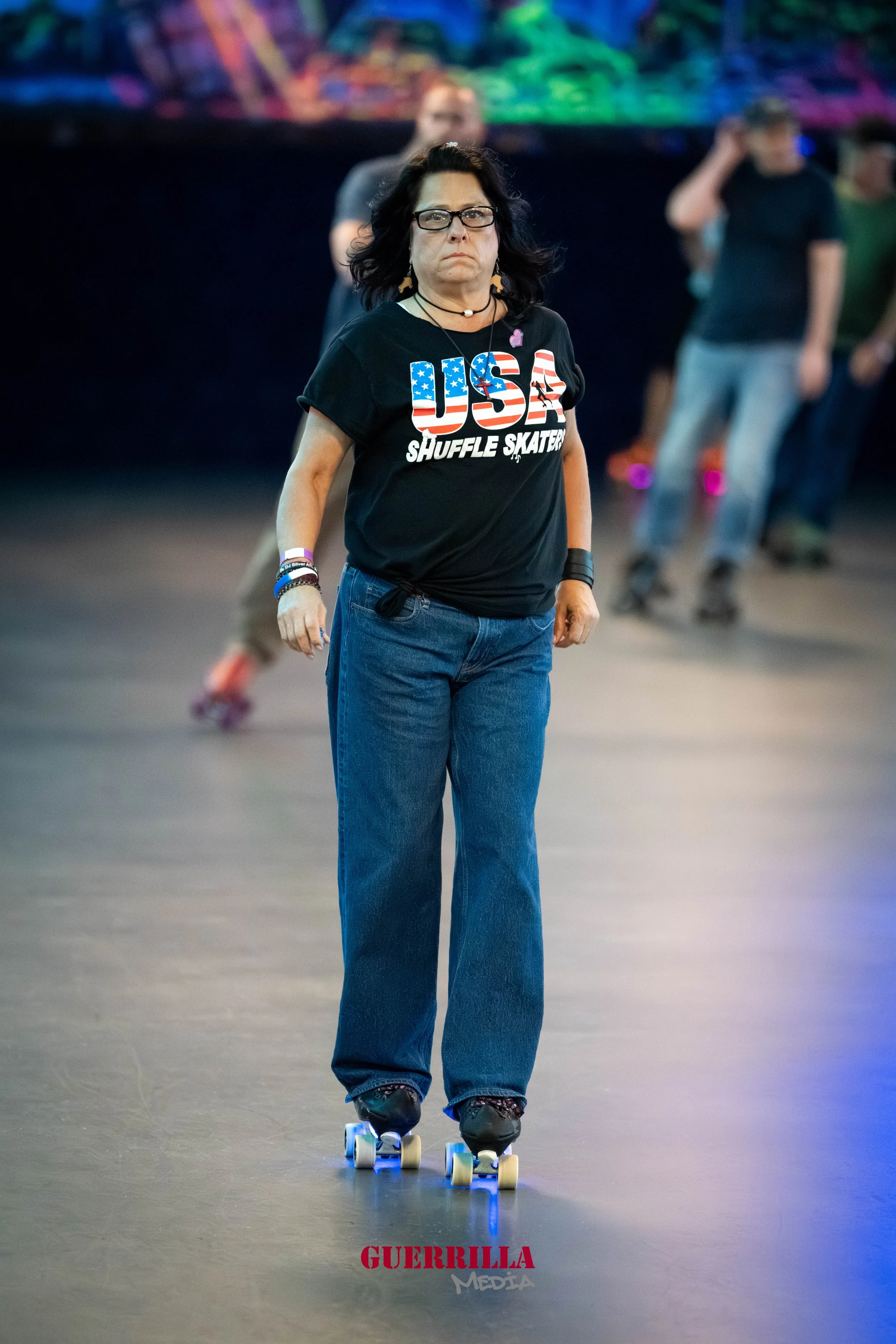 A woman with dark hair, glasses, and a serious expression roller skating indoors. She wears a black T-shirt with 'USA Shuffle Skaters' and American flag design, blue jeans, and various bracelets.