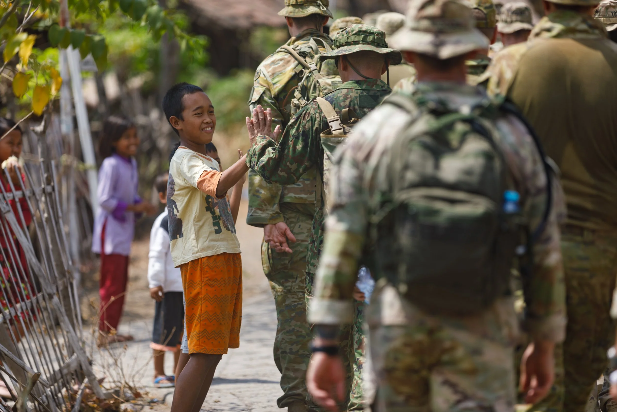 A young boy smiling and giving a high five to a soldier in camouflage uniform during a military outreach or community engagement, with other children in line and trees in the background.