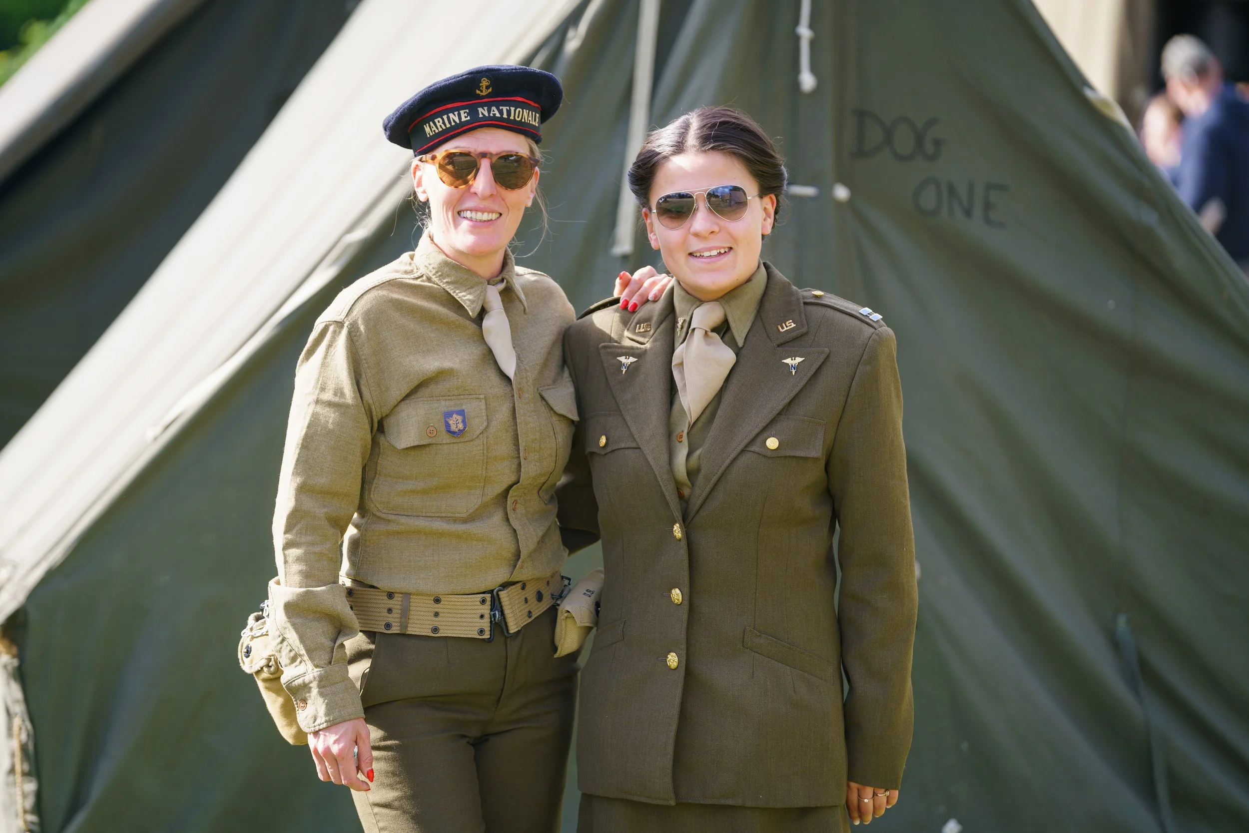 Two women in military uniforms smiling, standing outdoors in front of a green tent marked 'Dog One'.