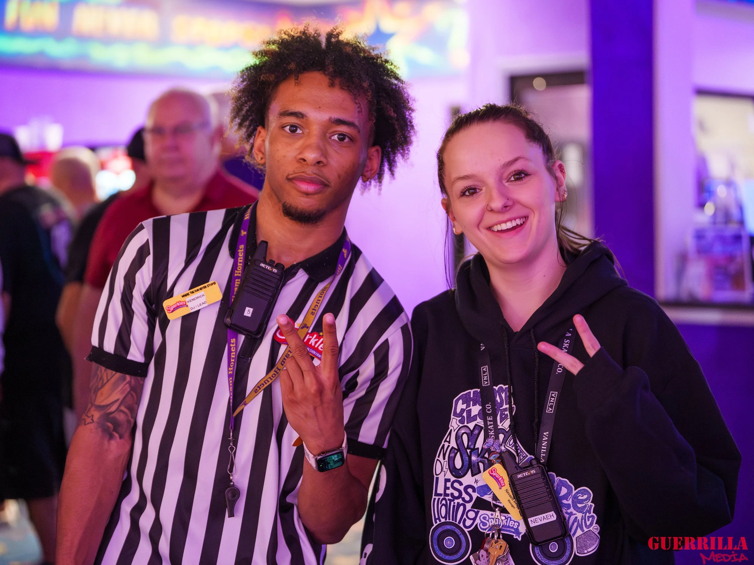 A young man and woman are standing together and smiling at the camera in an arcade. They are making a peace sign with their hands. The man is wearing a black and white striped referee shirt with a lanyard and badge, and the woman is wearing a black h