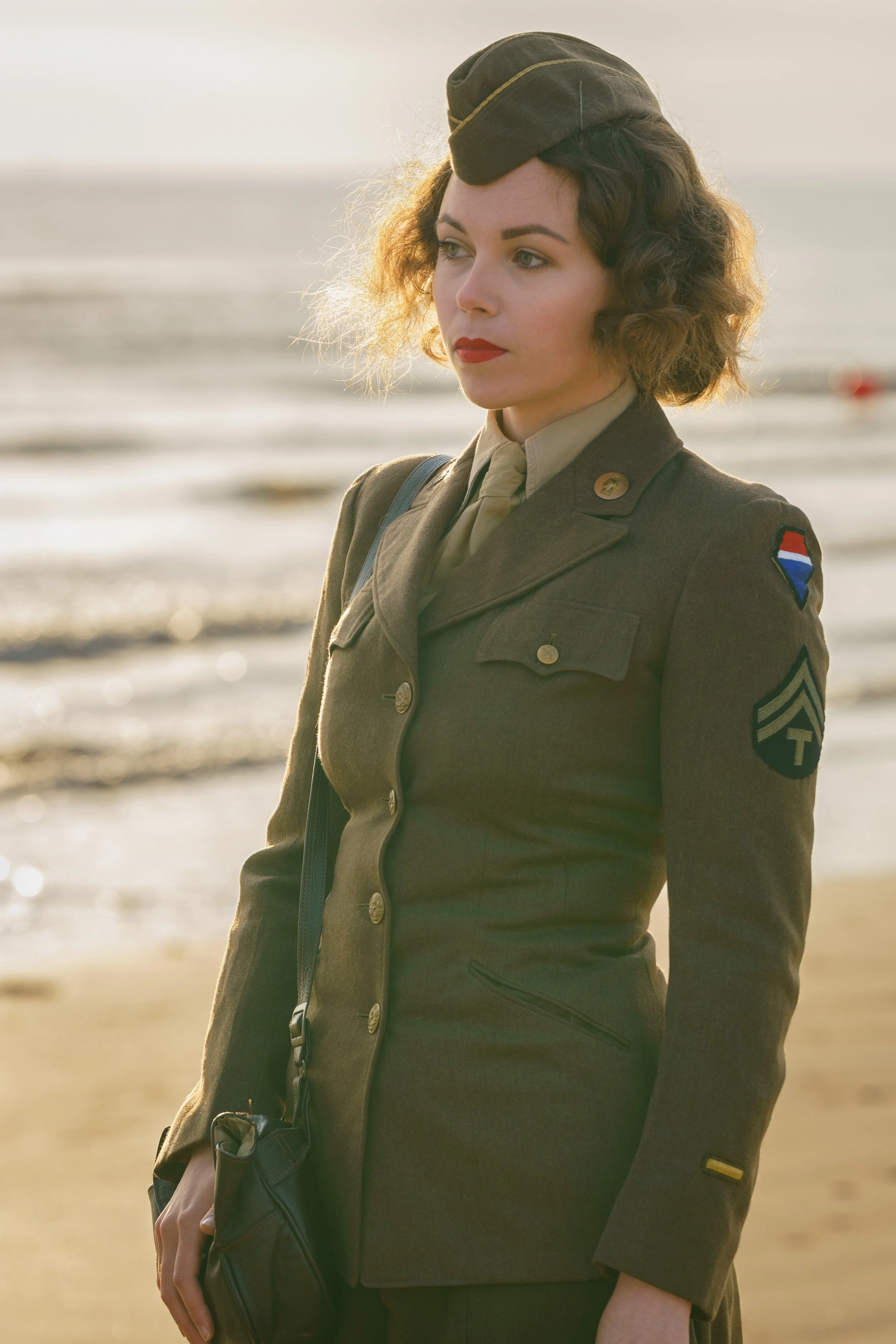 A woman dressed in a military uniform, standing on a beach with the ocean in the background.