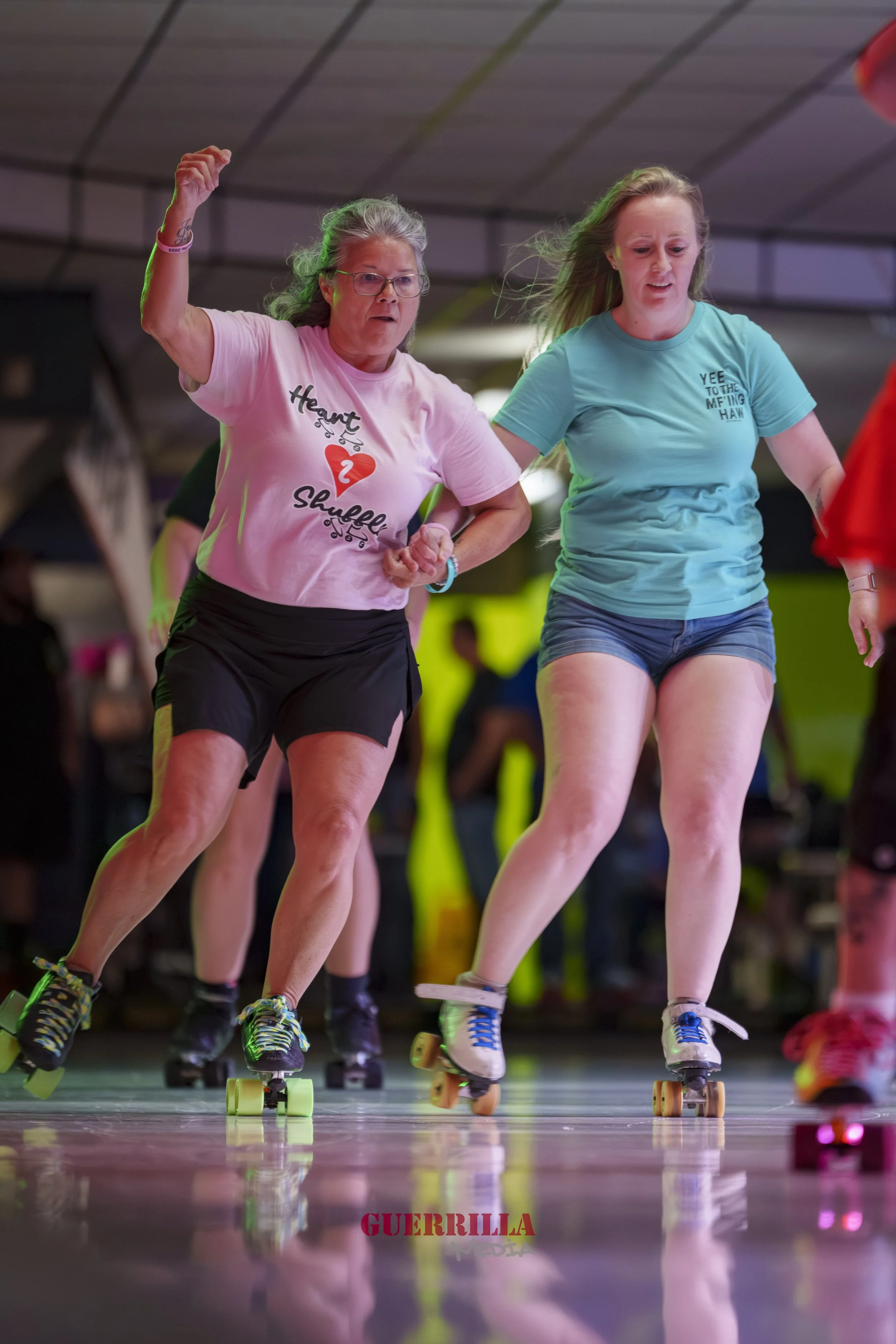 Two women roller skating indoors, one with gray hair and glasses wearing a pink shirt, the other with blonde hair wearing a teal shirt. They are holding hands and skating together.