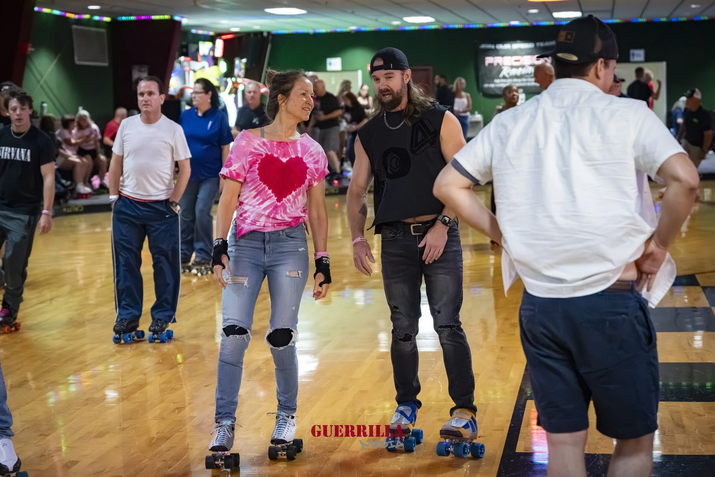 People roller skating at an indoor rink, some talking and socializing, colorful lights overhead, and a green wall with a movie poster in the background.