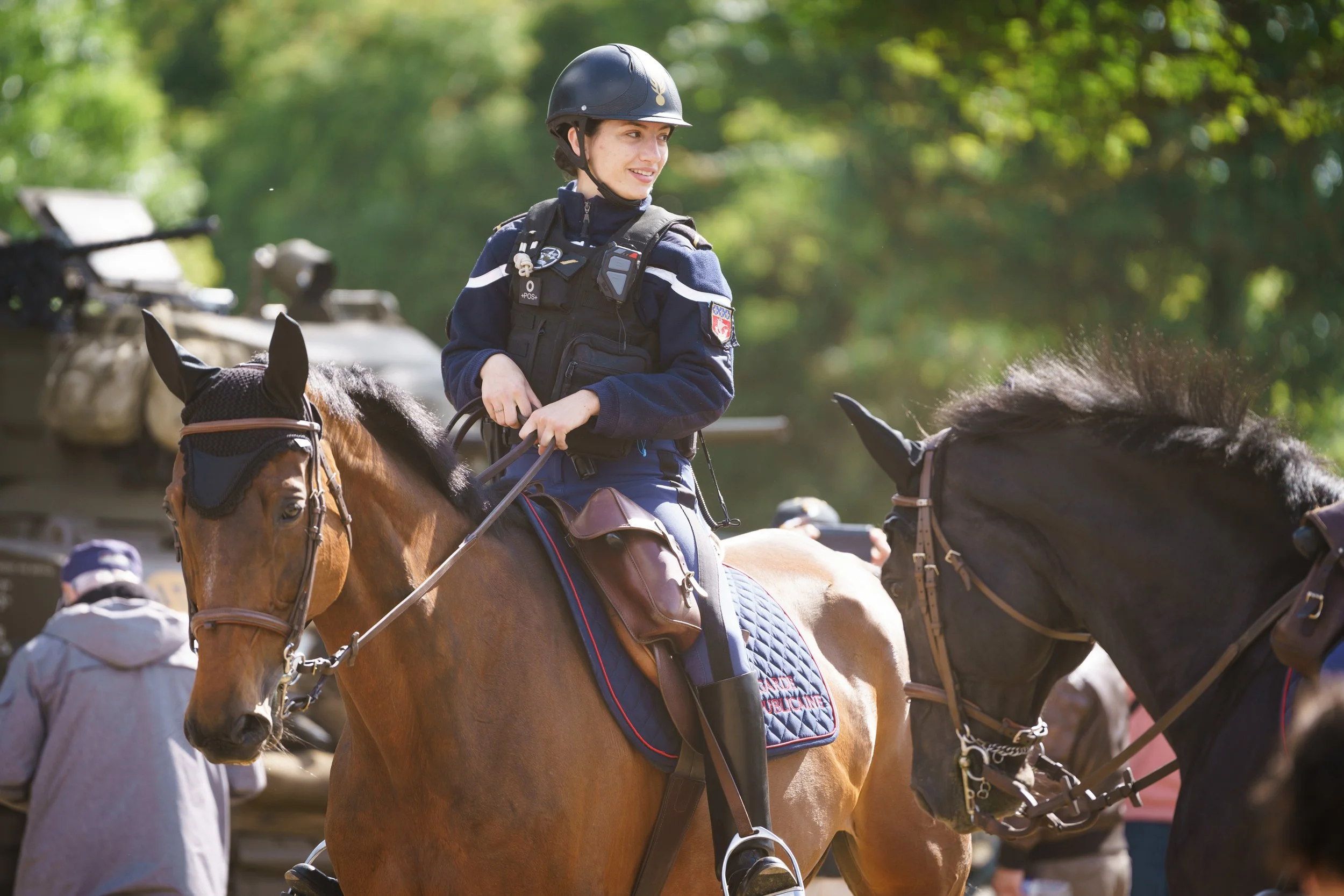 A young girl in a police uniform riding a horse, smiling, with green trees in the background.