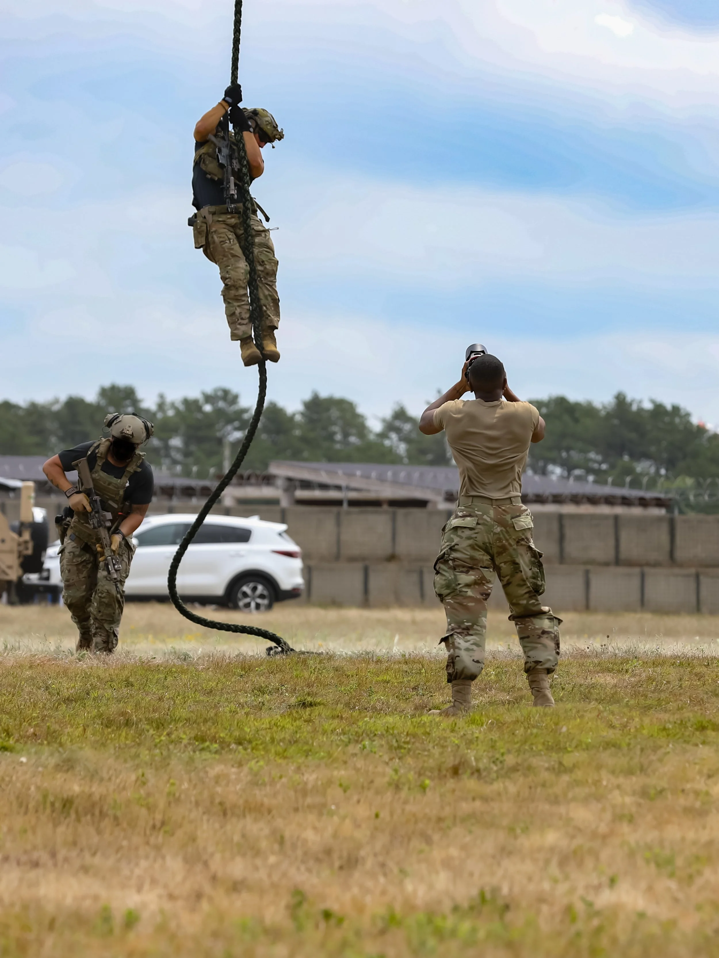 Military personnel practicing rope climbing and photography outdoors in a field with a white vehicle, trees, and a fence in the background.