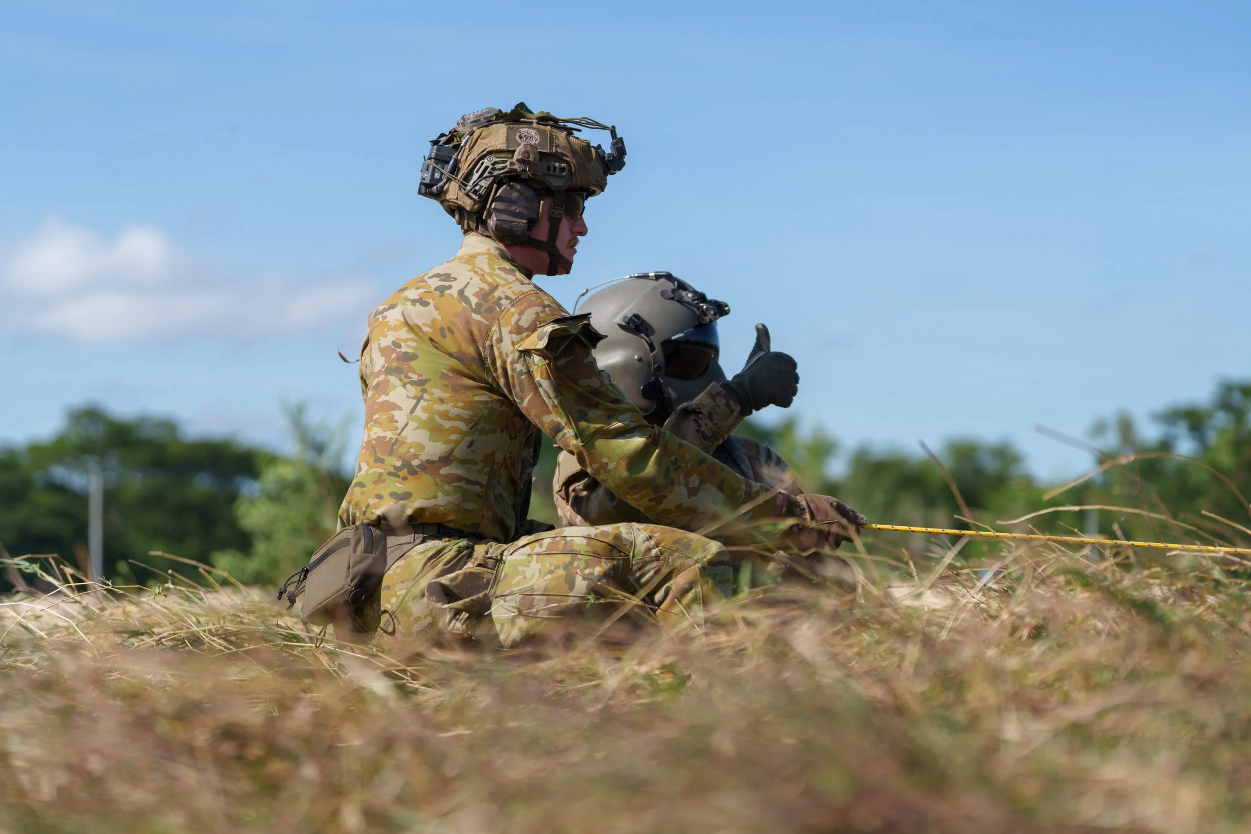 Two soldiers in camouflage uniforms kneel on a grassy field, one holding a helmet and giving a thumbs-up gesture, with a clear blue sky and trees in the background.