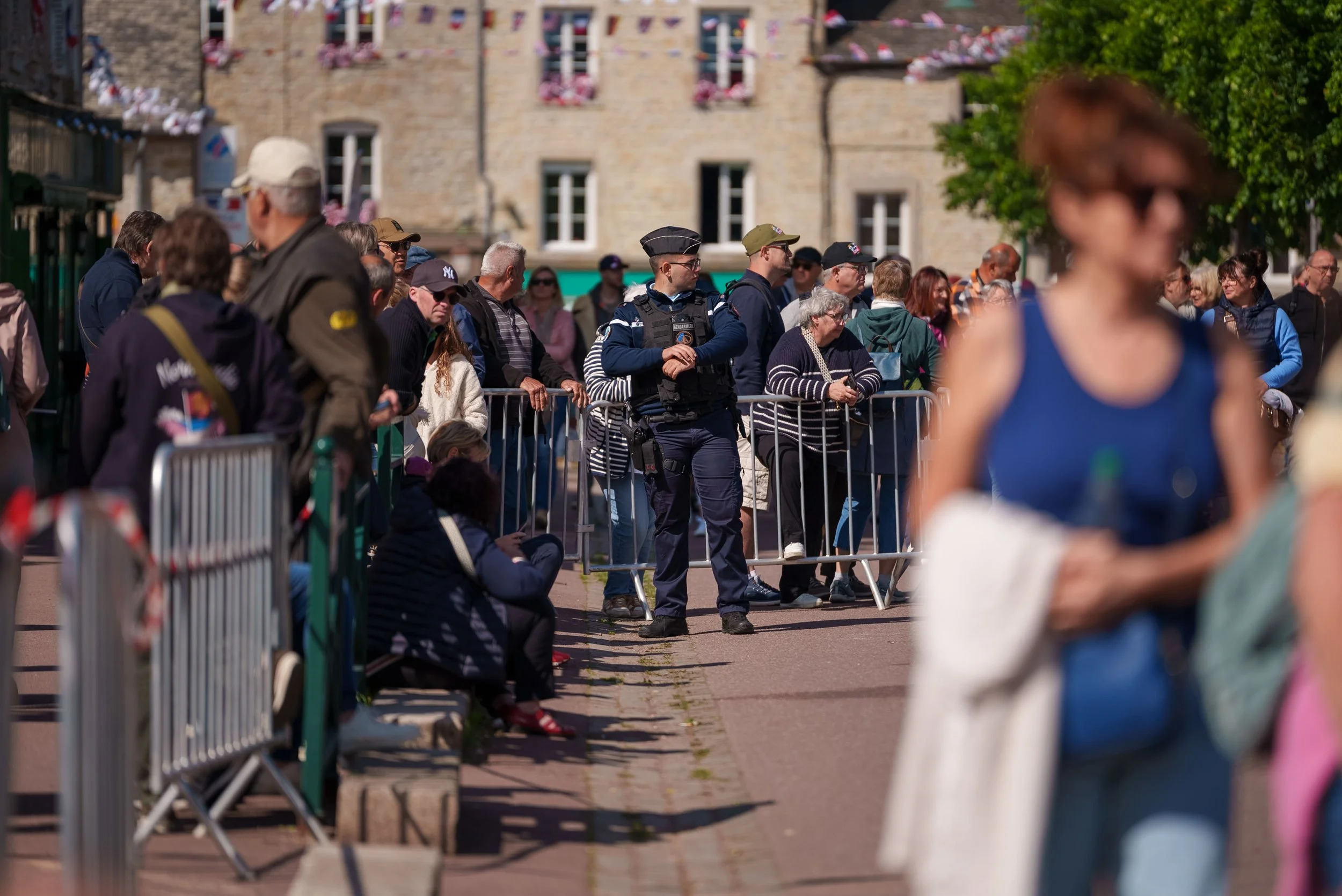 People gathered behind a metal barricade in an outdoor setting with a historic building in the background, some officials in uniform, and a woman in sunglasses in the foreground.