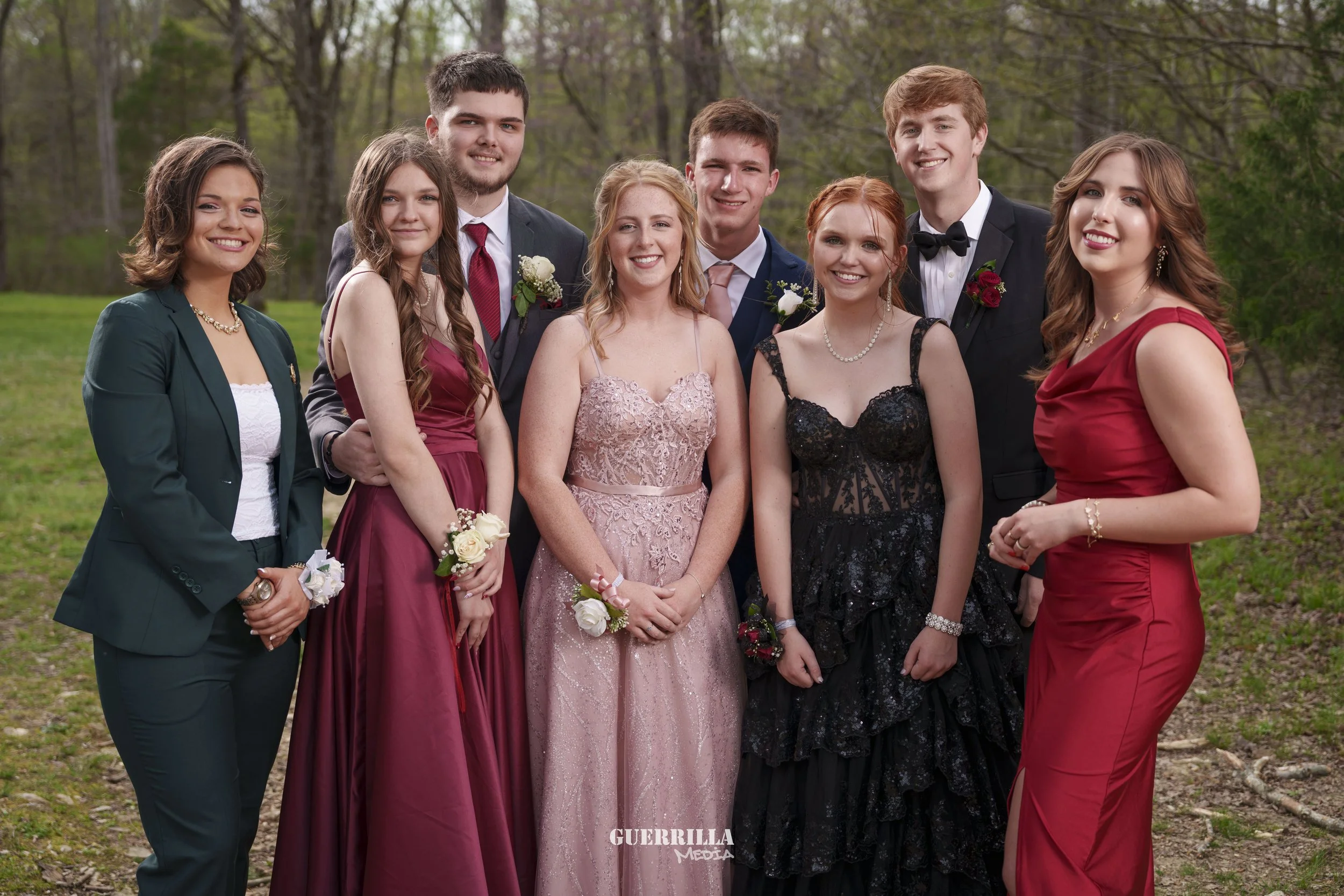 Group of nine young people dressed in formal attire standing outdoors in a park-like setting