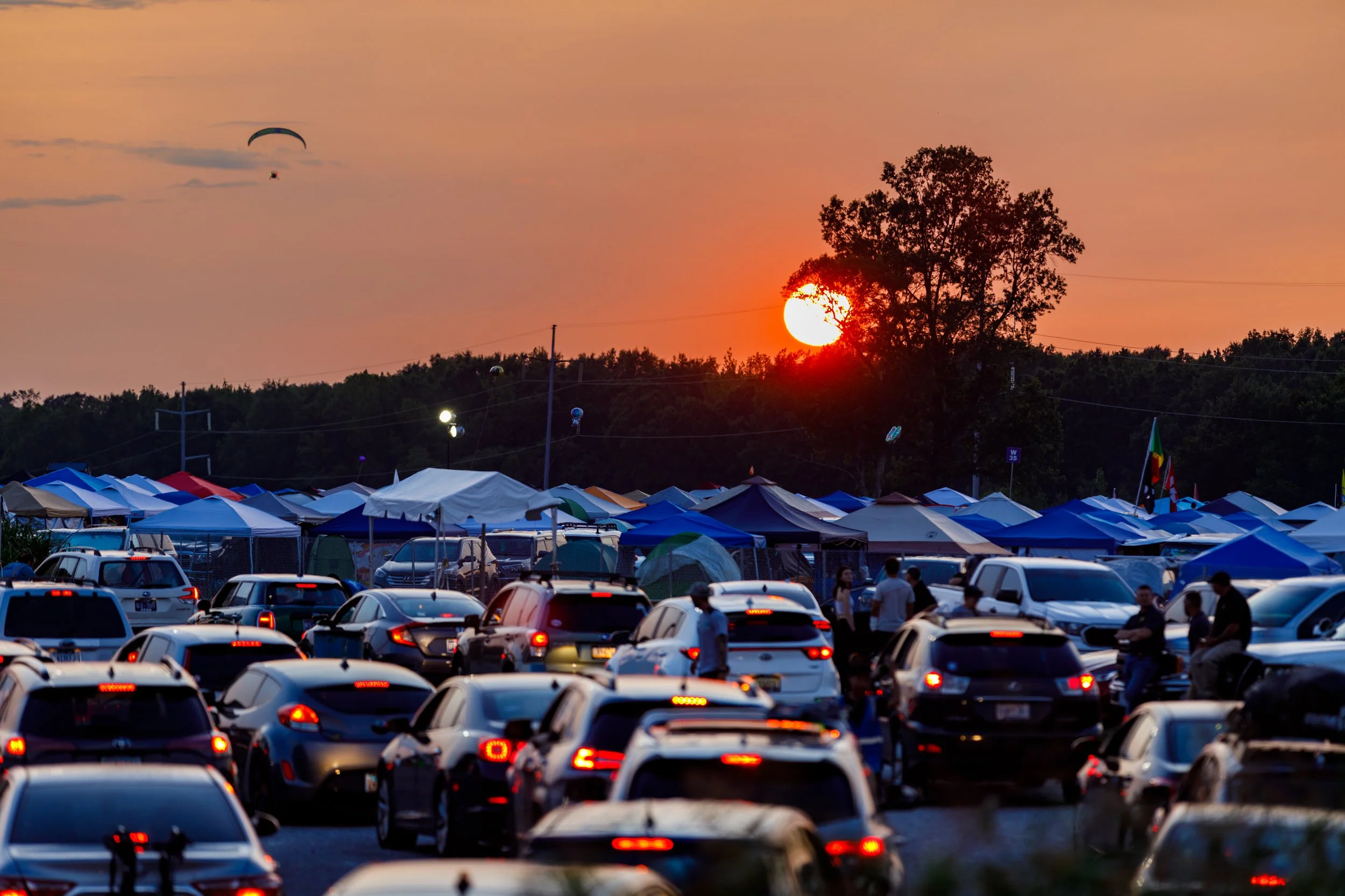 Scene of a parking lot filled with cars, tents, and people during sunset with the sun near the horizon, behind trees, and a paraglider flying in the sky.
