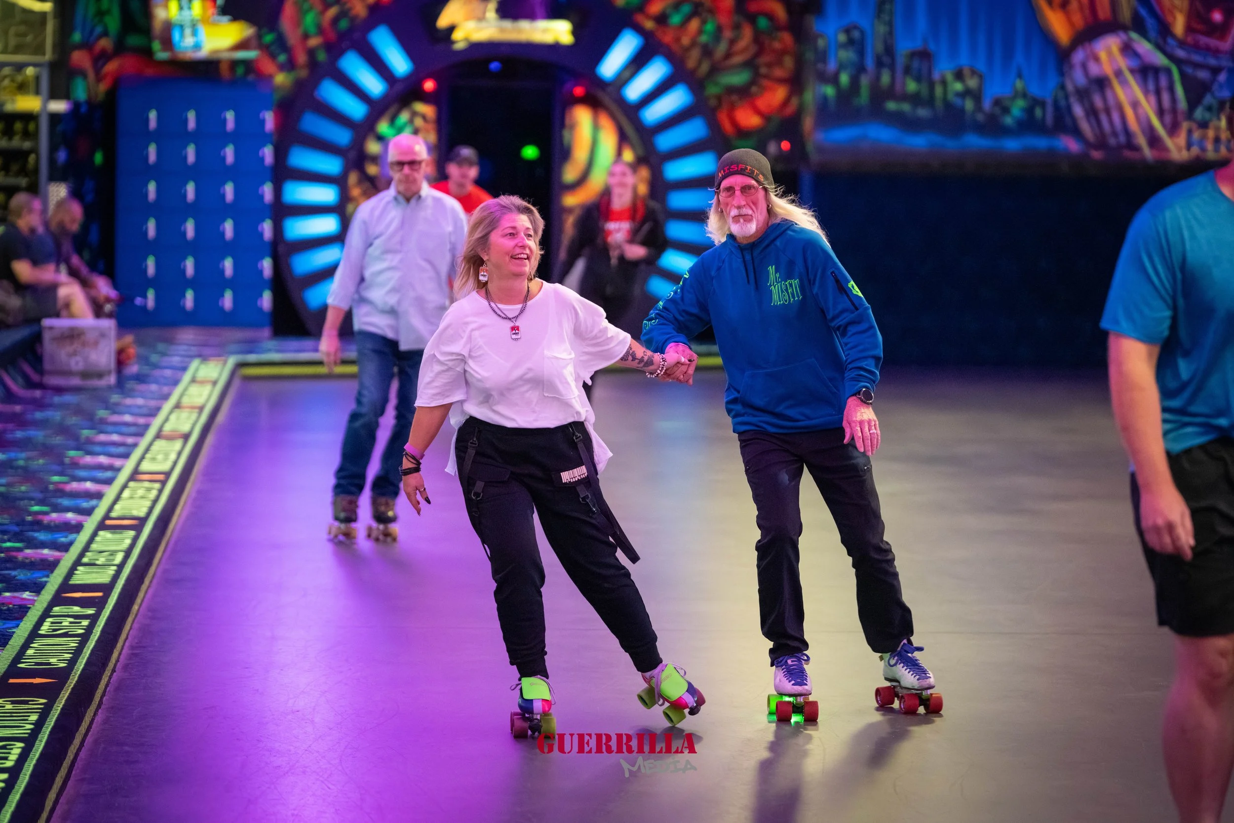Two people roller skating, holding hands and smiling, inside a colorful roller rink with neon lights. Other skaters and onlookers are visible in the background.