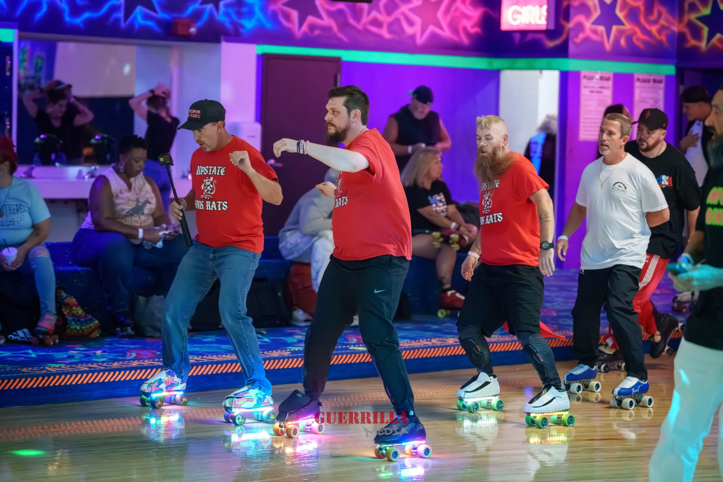 People roller skating at an indoor roller rink with colorful, neon lighting and a lively atmosphere. Some skaters are wearing red shirts with the text 'Lipstick and Skates'.