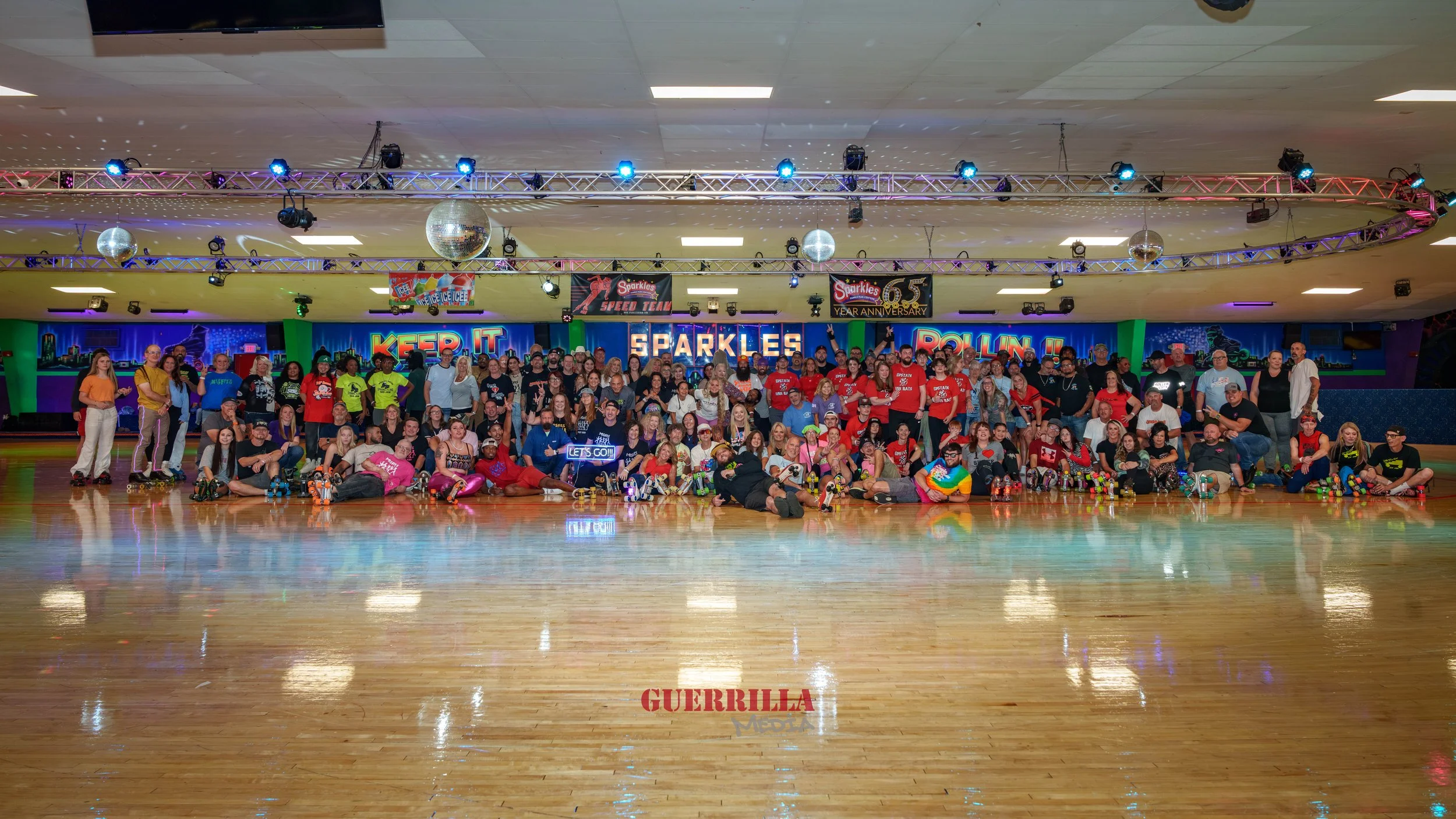 Large group of people at an indoor roller skating rink with neon lights and disco balls, posing for a group photo. The background features colorful signs and decorations celebrating an anniversary.