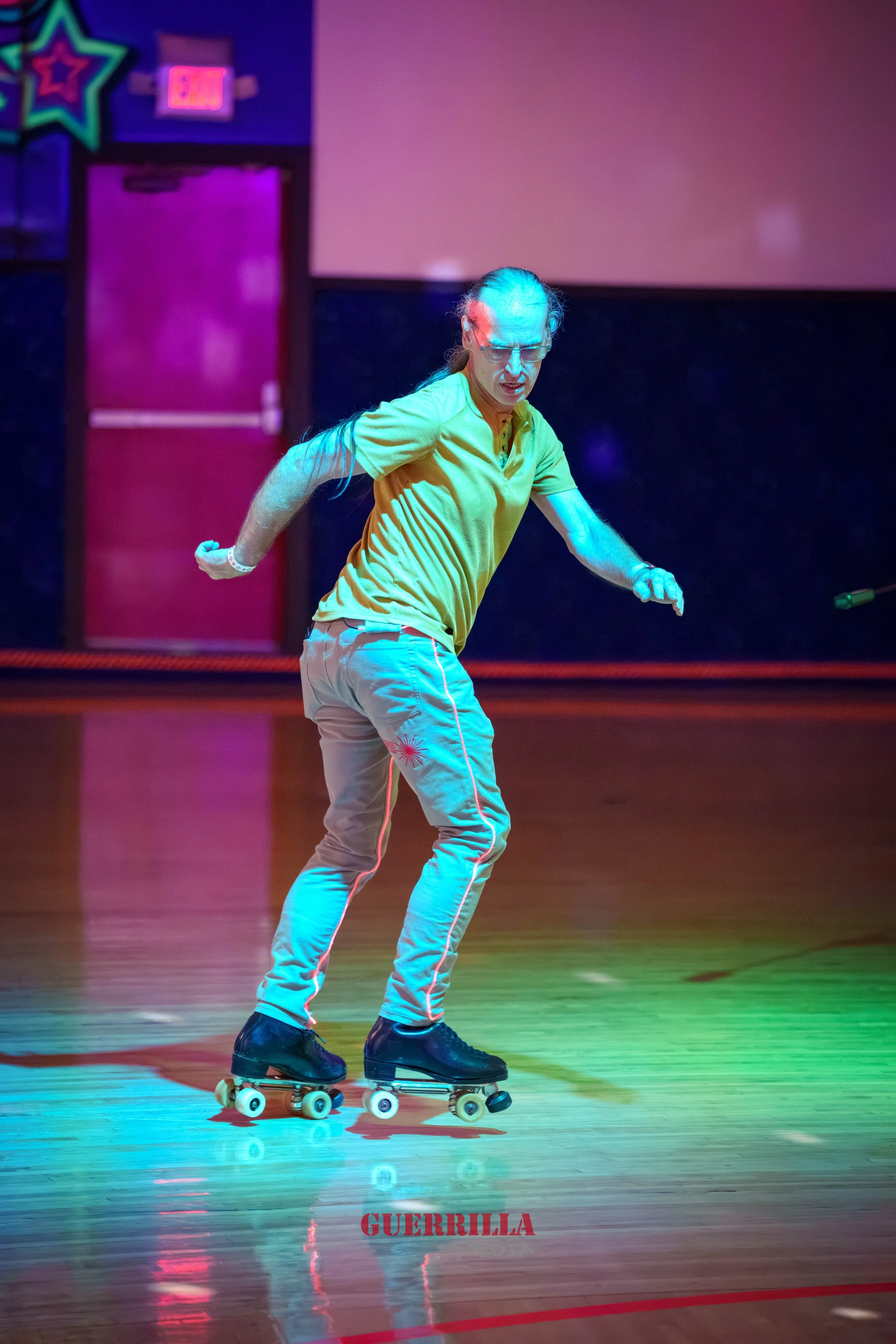 A man with long hair, glasses, wearing a yellow shirt and light pants, rollerskating on a wooden floor in a colorful, neon-lit environment. The word 'GUERRILLA' is written in red at the bottom of the image.