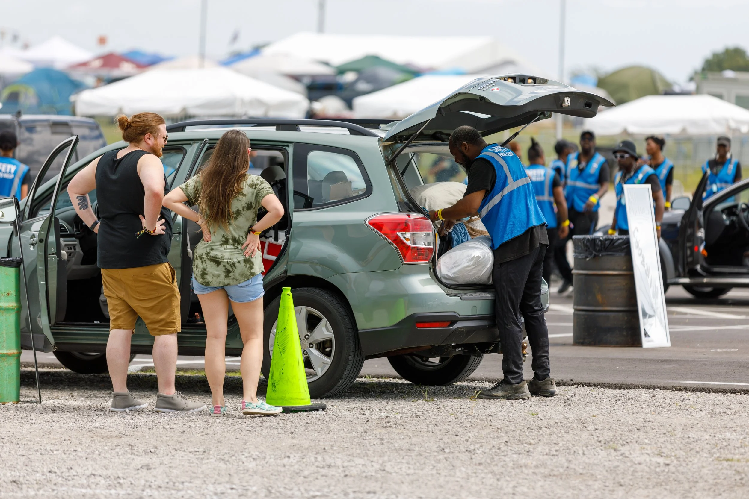 Two women and a man standing near the open trunk of a green SUV at an outdoor event, with security personnel and tents in the background.