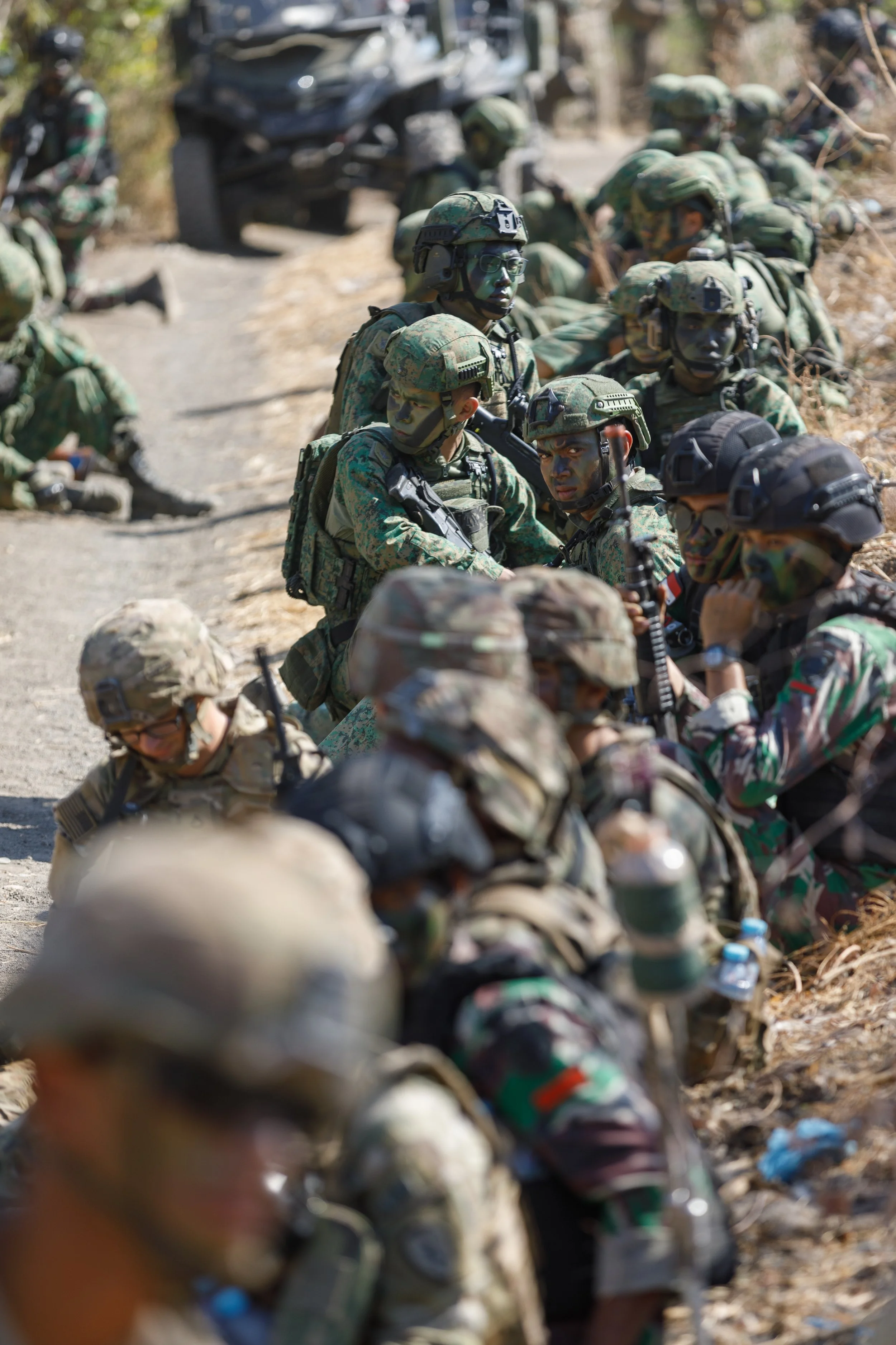 Soldiers in camouflage uniforms sitting and kneeling on the ground outdoors, some with face paint, gear, and helmets, during a military event or training.