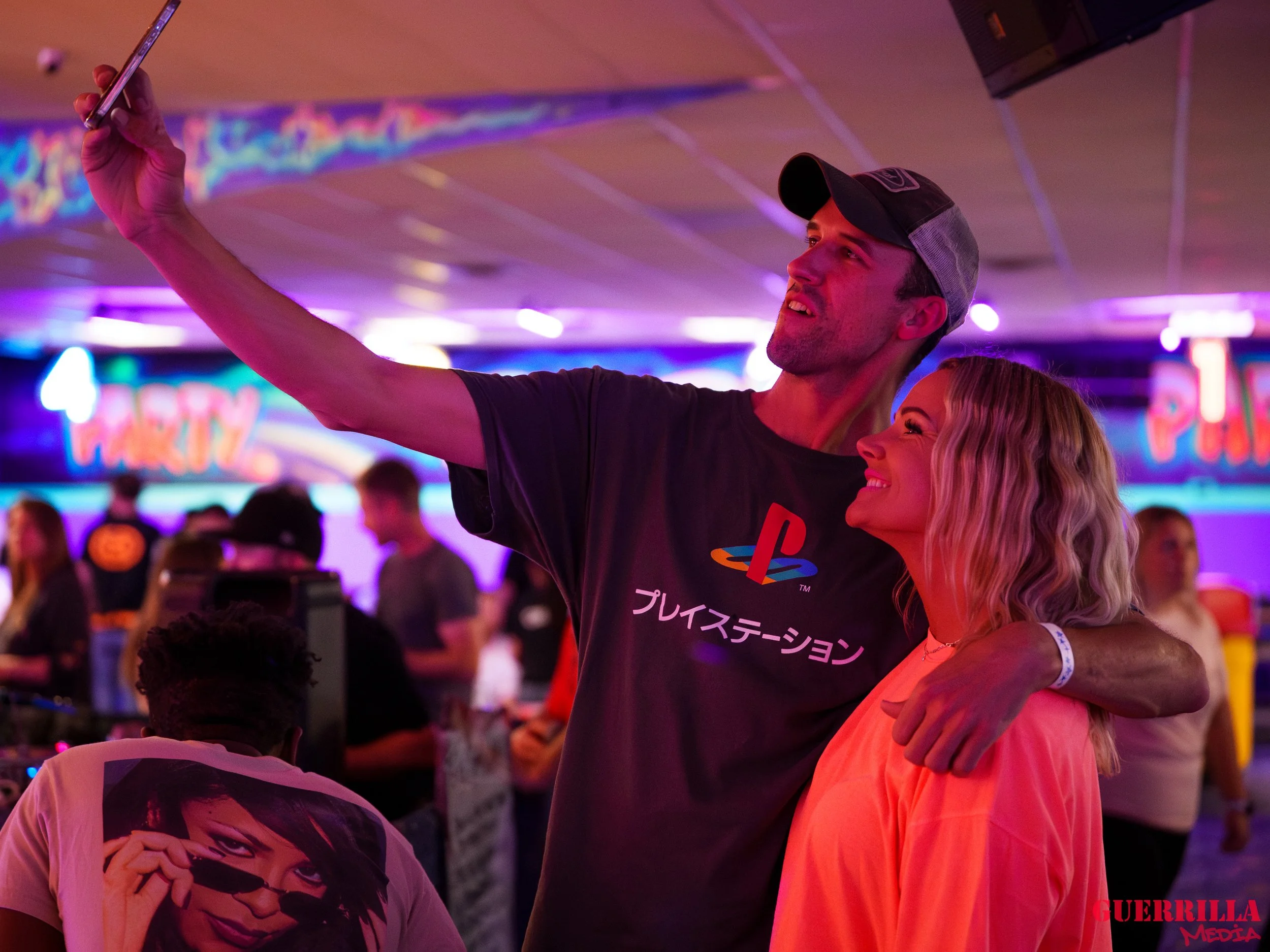 A man and woman taking a selfie together at a party. The man is wearing a black T-shirt with a PlayStation logo and Japanese text, and a baseball cap. The woman has blonde hair and is smiling. The background shows a lively party scene with colorful n
