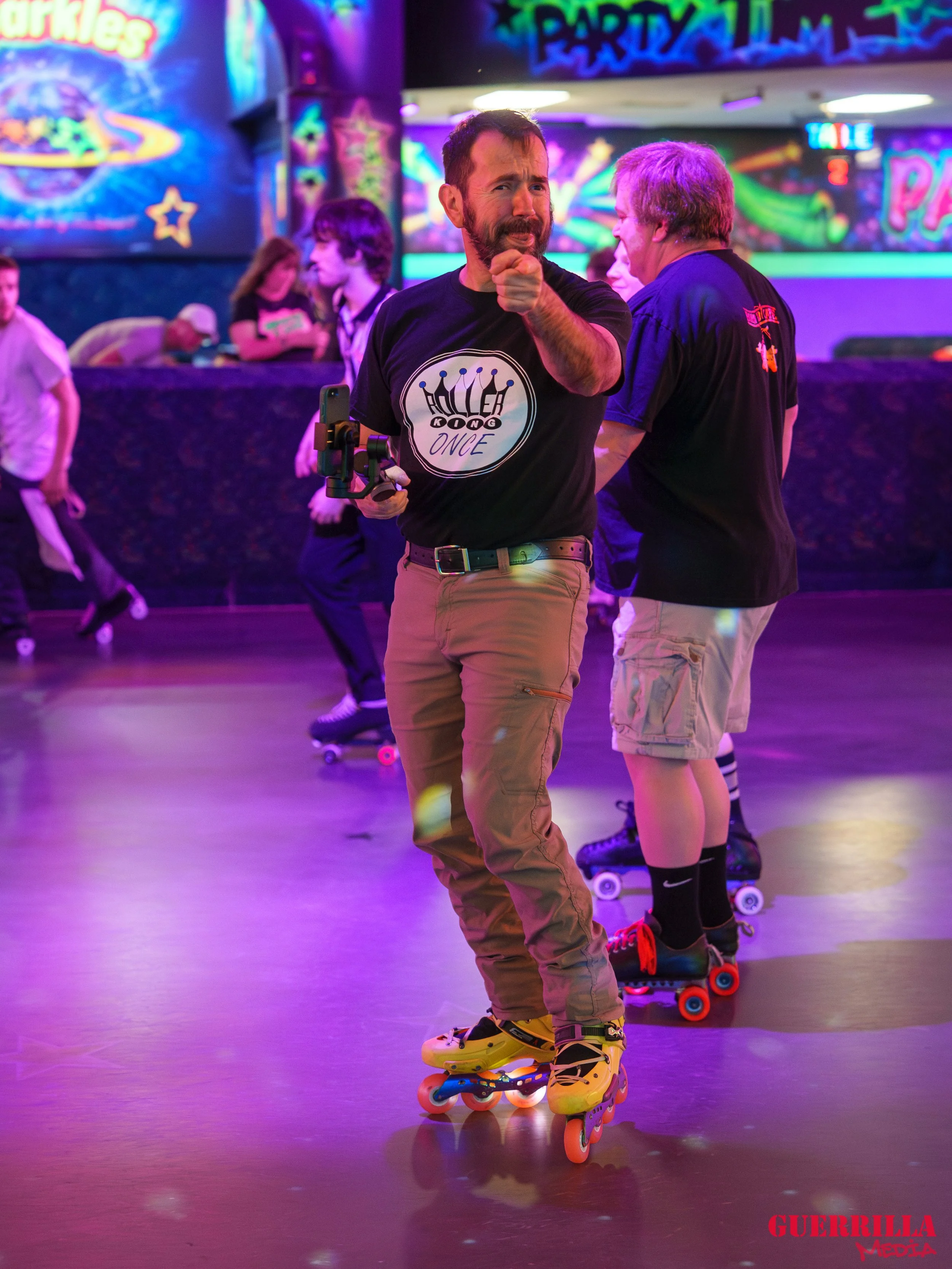 A man roller skating at an indoor roller rink with neon lights, pointing at the camera, surrounded by other skaters, with colorful wall decorations and a sign that says 'Party' in the background.