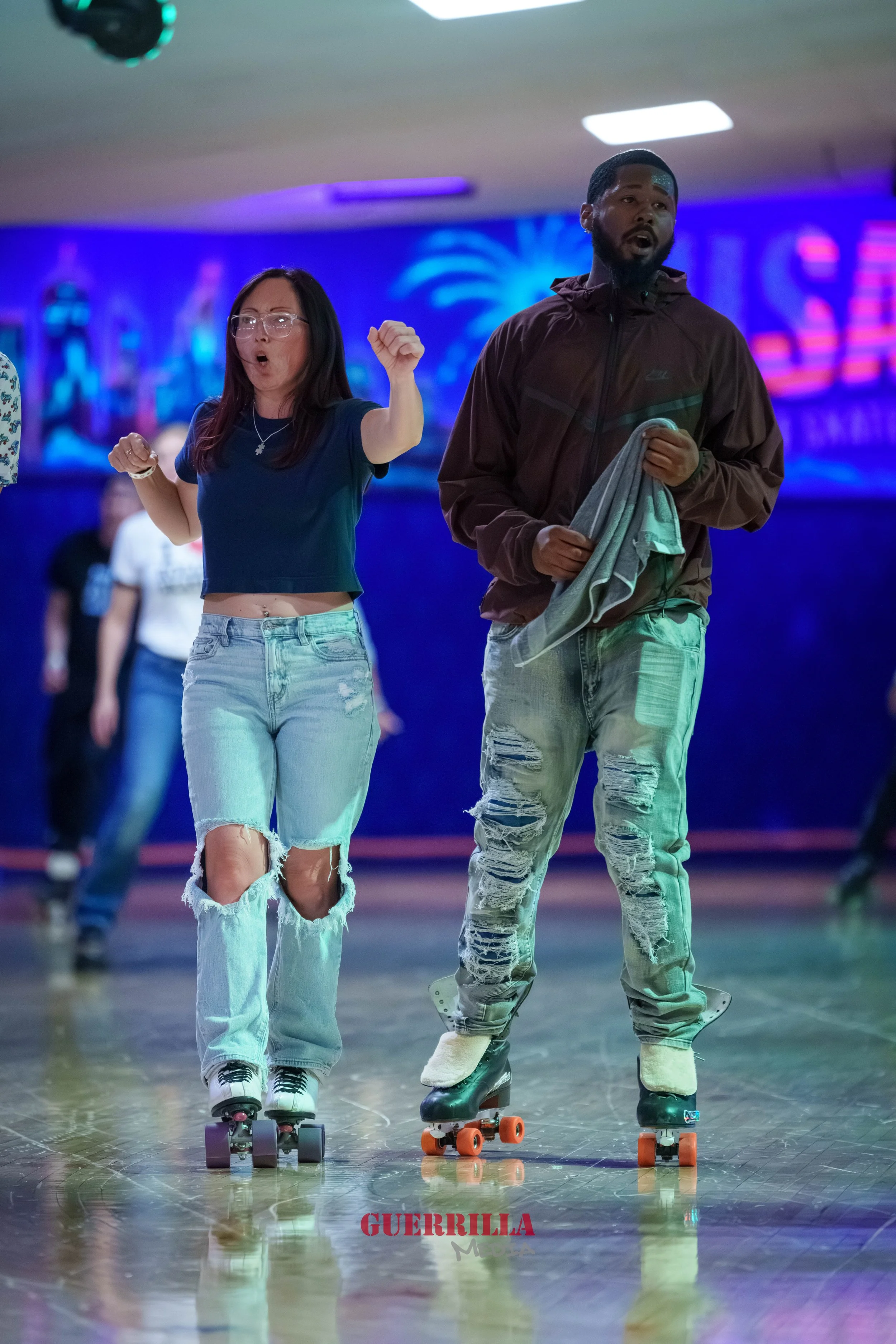 Two people roller skating indoors, one woman and one man, with colorful neon lights in the background.