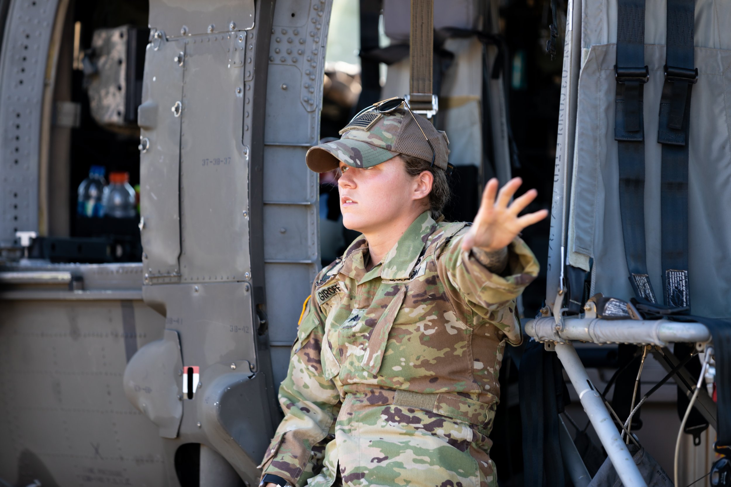 A woman in military uniform with a camouflage pattern, wearing a cap with a flag patch, is gesturing with her hand while sitting next to military equipment and an aircraft.