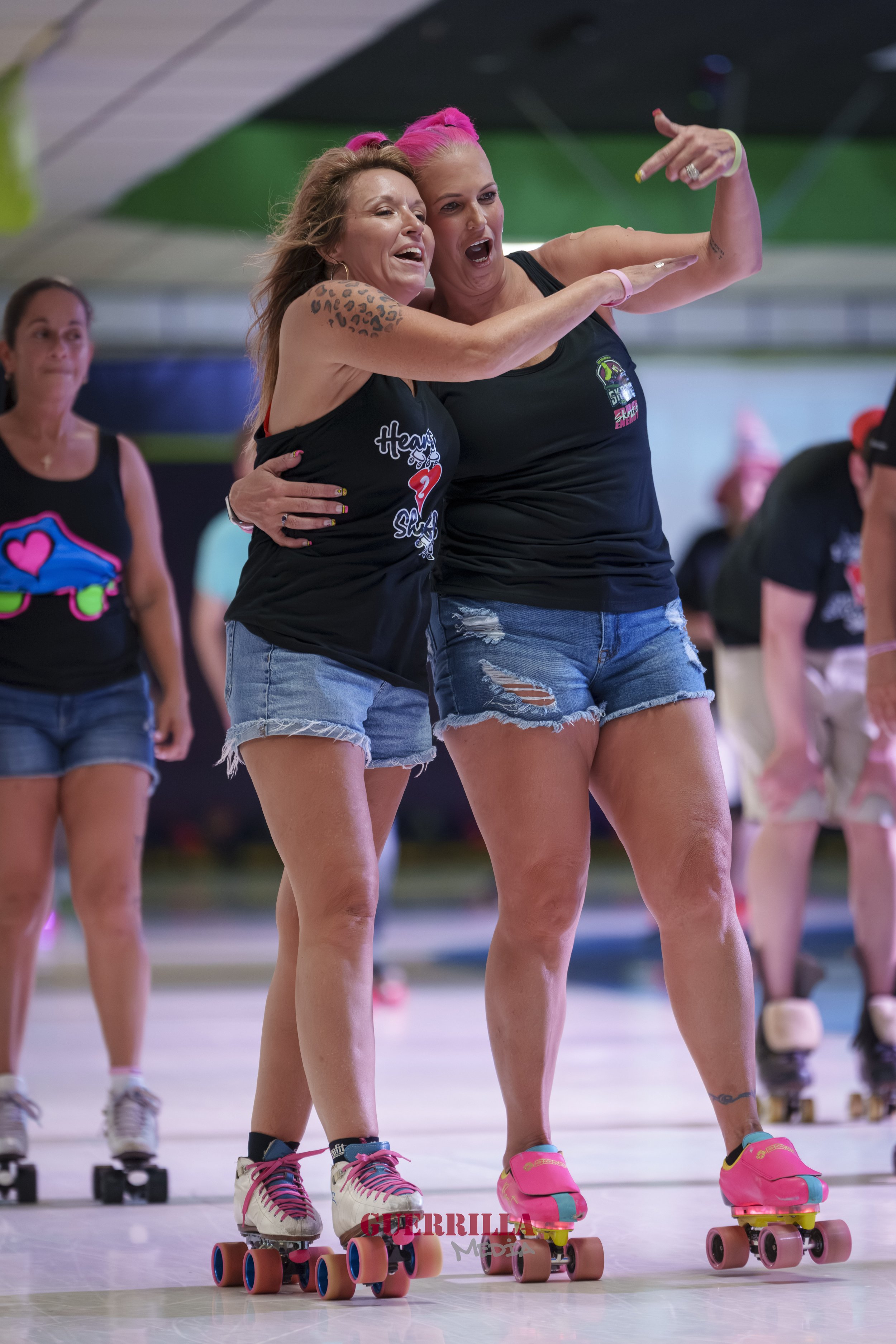 Two women roller skating together, dance or celebrate, with other women in the background, wearing casual summer clothing indoors.
